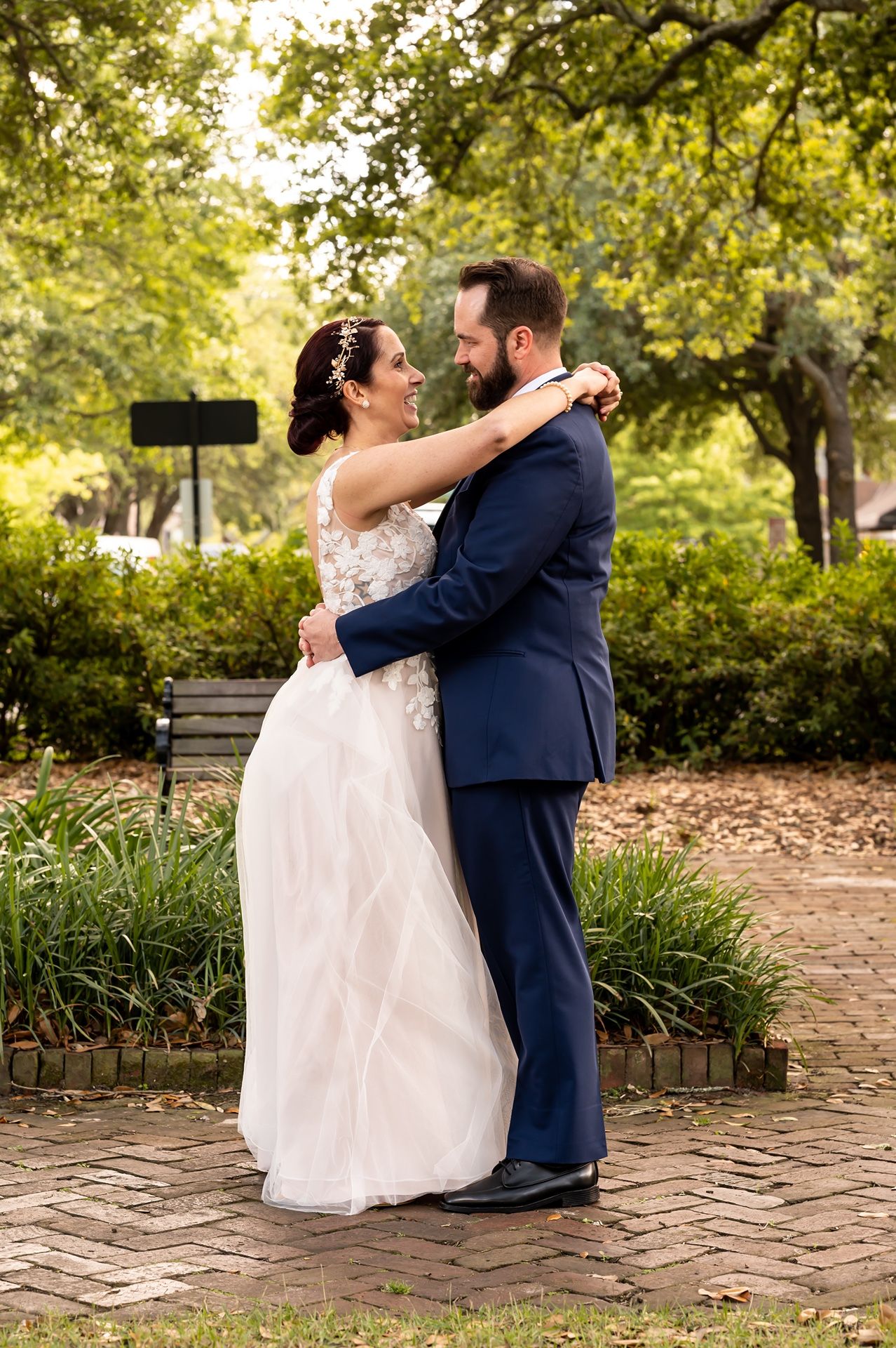 A couple embraces on a brick pathway in a lush garden setting during their wedding, wearing formal navy and white attire.