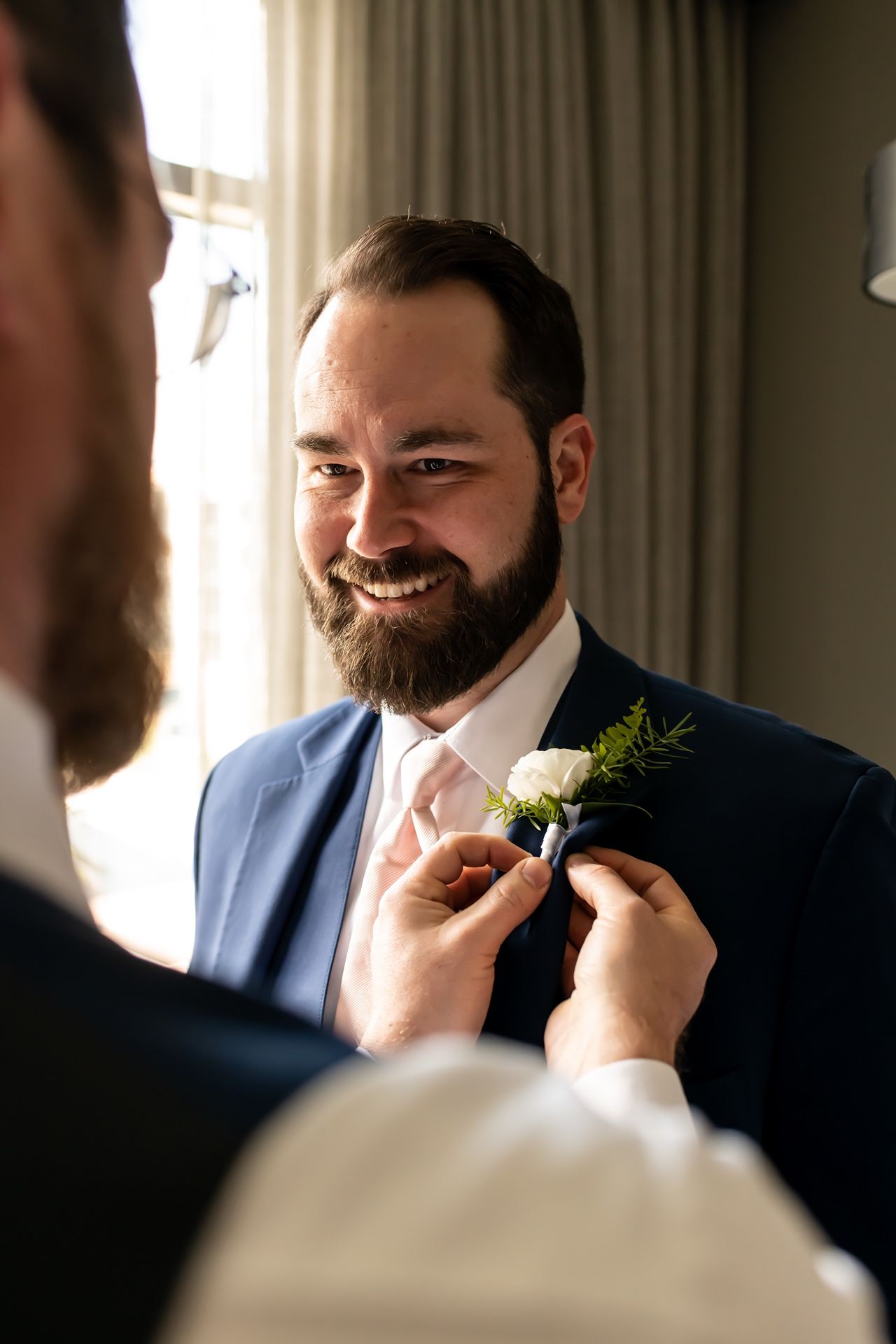 A groom in a blue suit gets help adjusting his floral boutonniere before the ceremony.