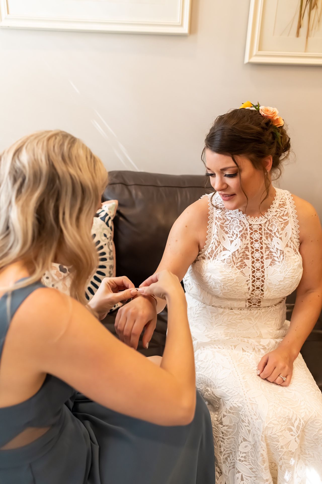 A person in an elegant lace wedding gown sits on a grey couch while another person helps with final preparations.