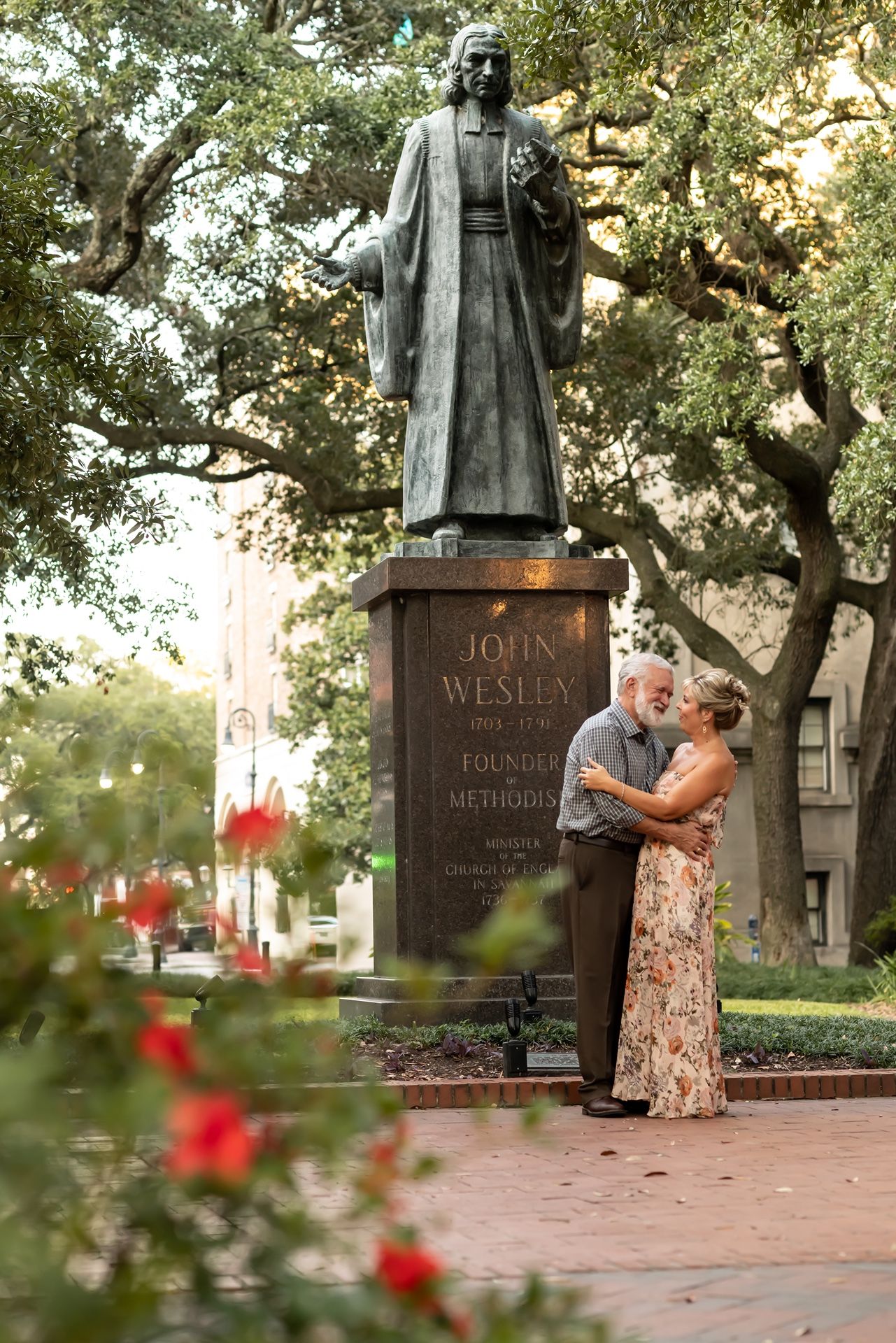 A couple embraces in front of the John Wesley statue surrounded by trees and red flowers.