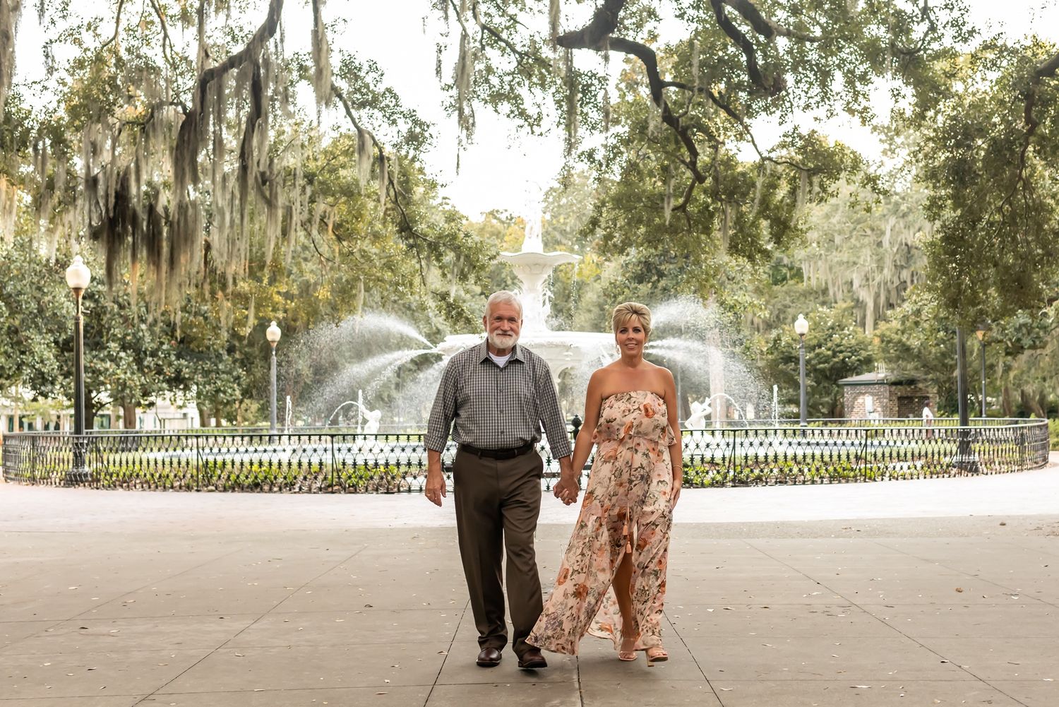 A couple takes a stroll through a park with Spanish moss trees and a fountain in the background.