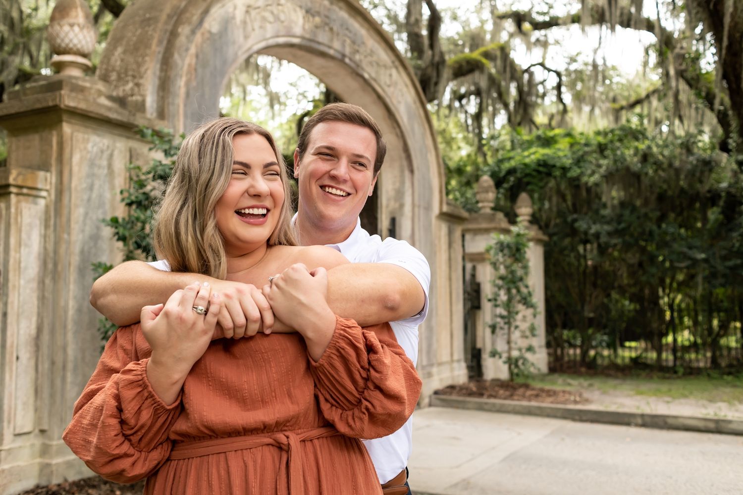 Couple embraces and laughs together under stone archway surrounded by Spanish moss and lush greenery.