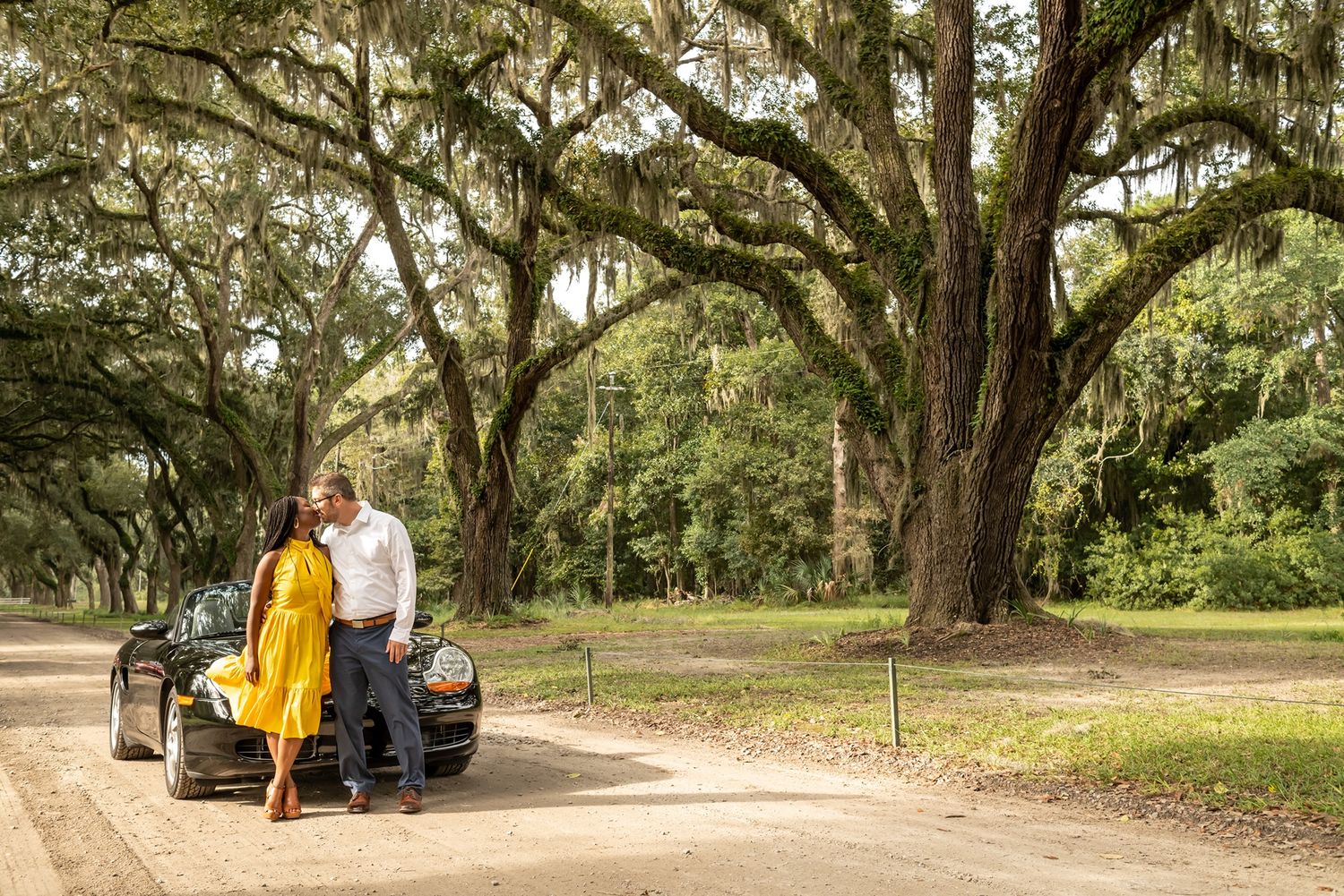 A couple stands by their vintage car beneath sprawling oak trees draped with Spanish moss on a dirt road.