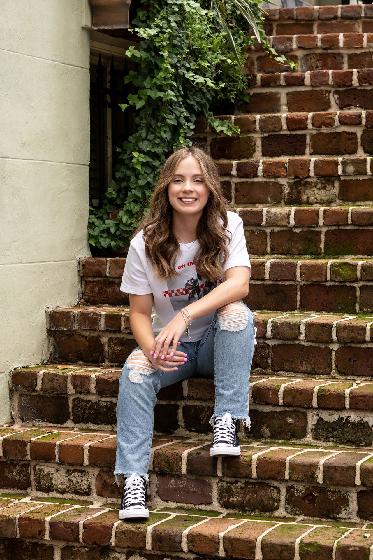 Person in jeans and white t-shirt sits casually on red brick steps with ivy-covered wall in the background.