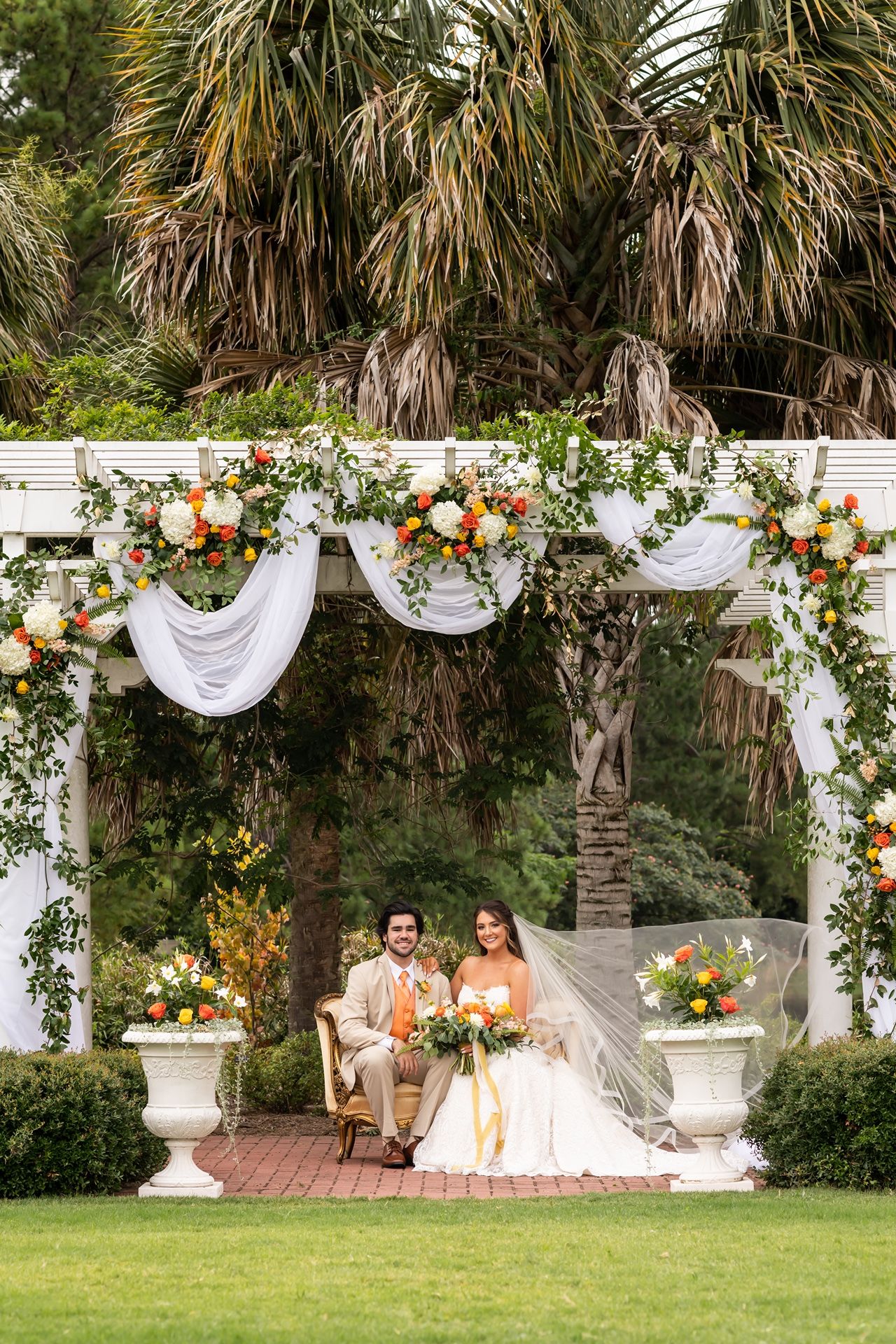 Elegant outdoor wedding ceremony setup with white pergola decorated in orange florals and greenery against tropical palm trees.