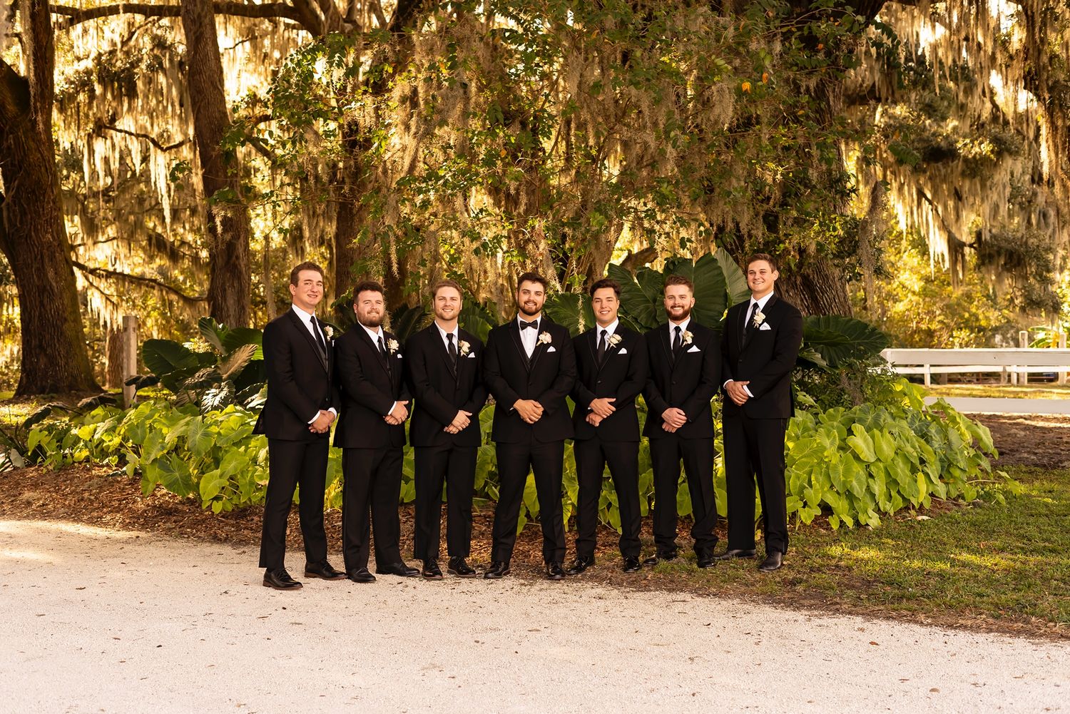 Large wedding party in formal attire poses outdoors against natural foliage and tree backdrop.