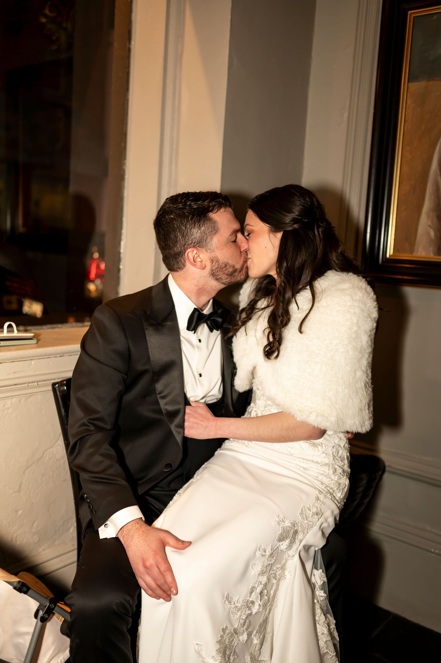 Newlywed couple share intimate kiss while sitting together in elegant formal attire at wedding reception.