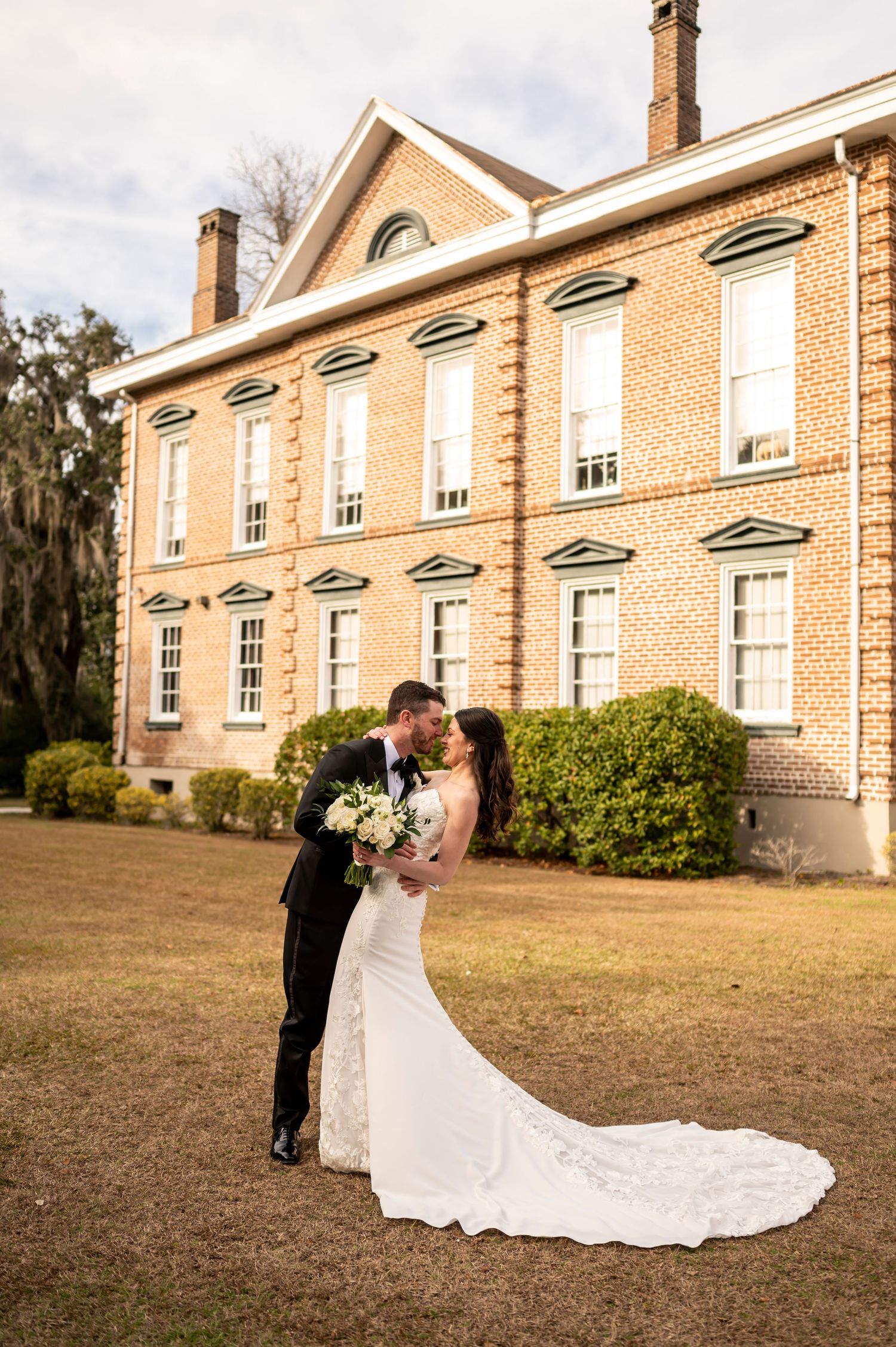 A romantic couple embraces outside a grand historic brick mansion with white trim and tall windows in a garden setting.