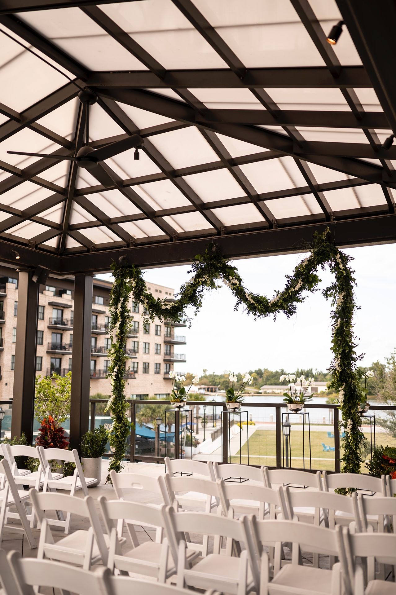 Wedding ceremony setup with white chairs under a covered pavilion decorated with floral garlands overlooking a marina.