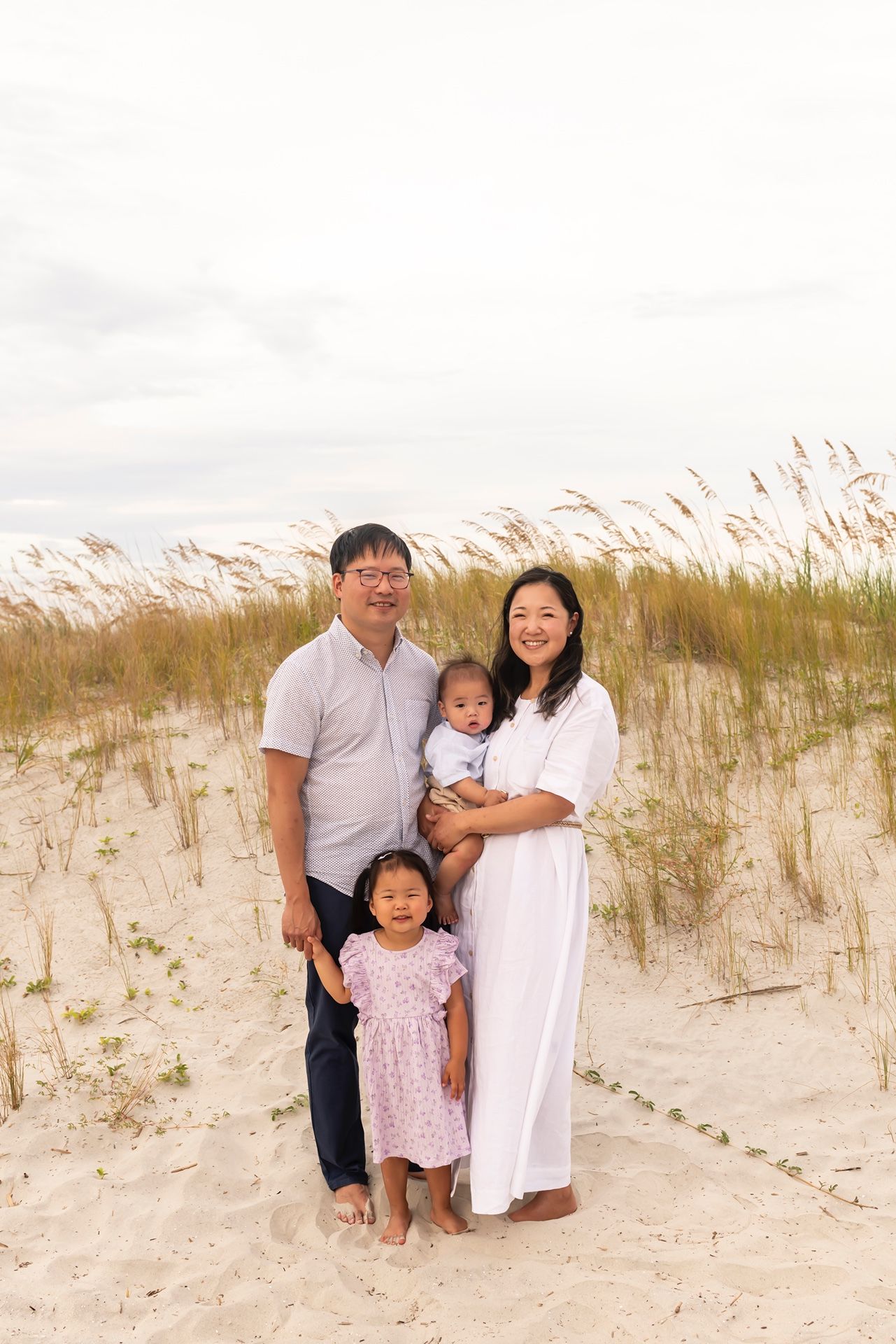 A family poses together on sandy dunes with sea grass waving in the background during sunset.