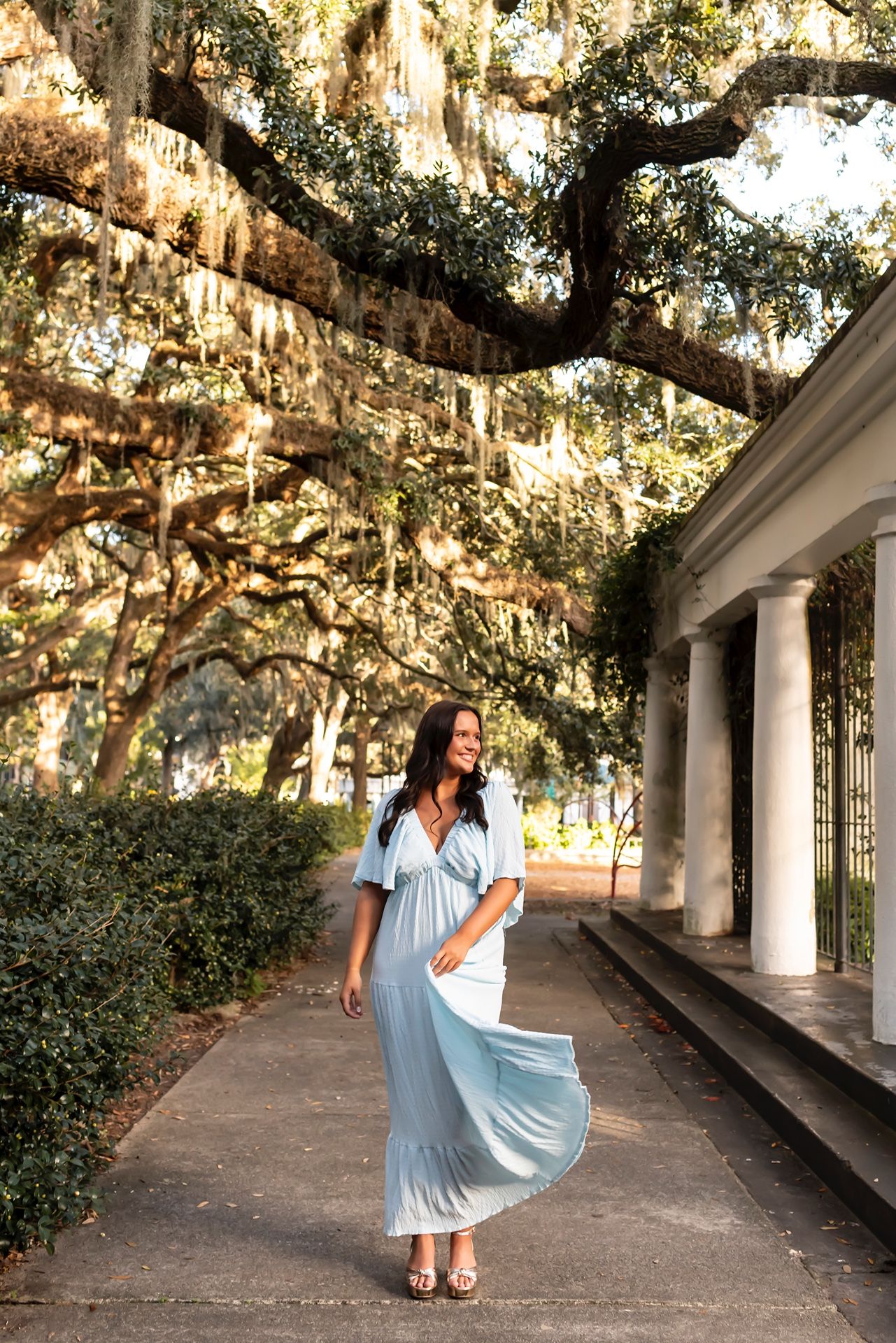 Woman in flowing light blue maxi dress walking along columned pathway under moss-draped oak trees in Southern setting.