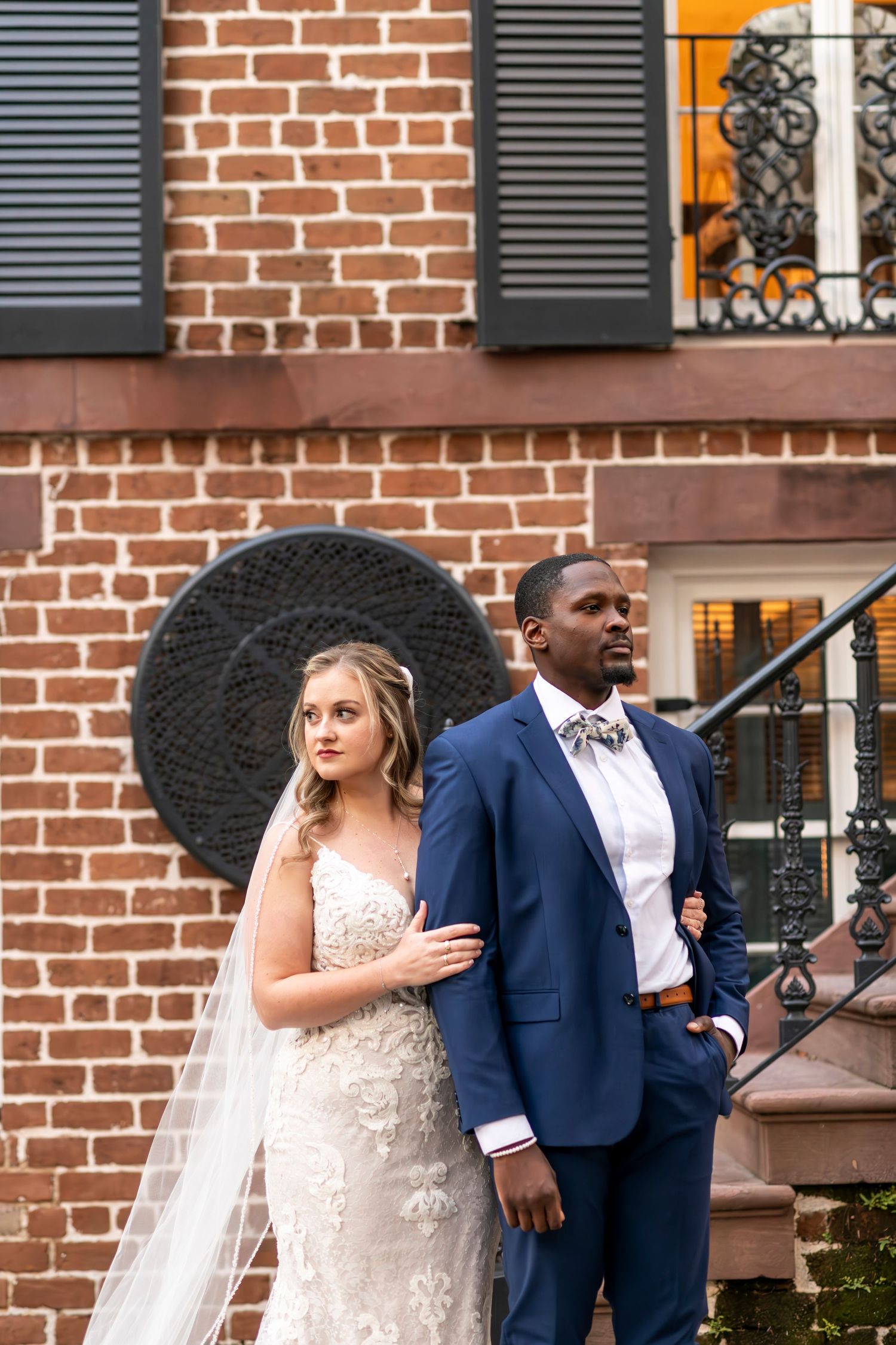 A couple in wedding attire stands together on brick steps of a historic building with black shutters and ornate railing.