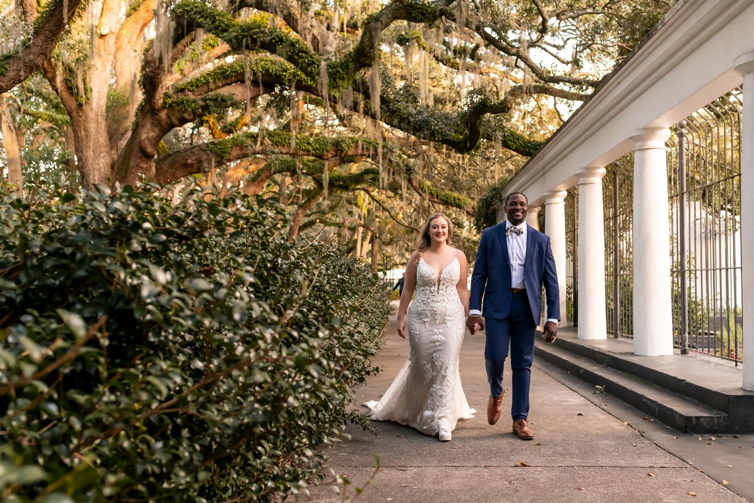 A couple walks along a white columned pathway under Spanish moss draped oak trees in Charleston, South Carolina.