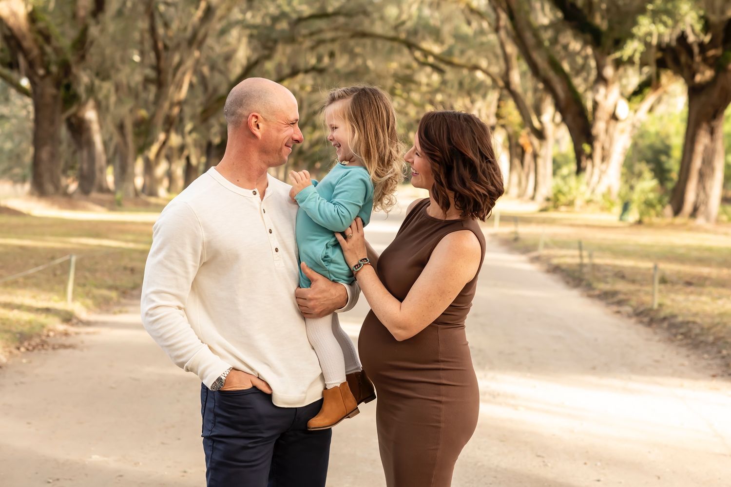 A couple holding a small child share tender moments together in a sun-dappled outdoor setting.