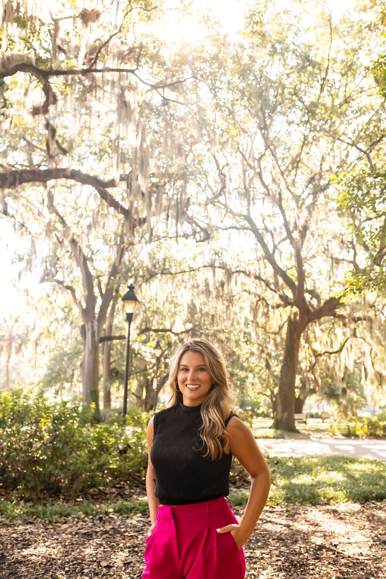 Person in a black sleeveless top and bright pink pants stands smiling in a sunlit park with Spanish moss draped trees.