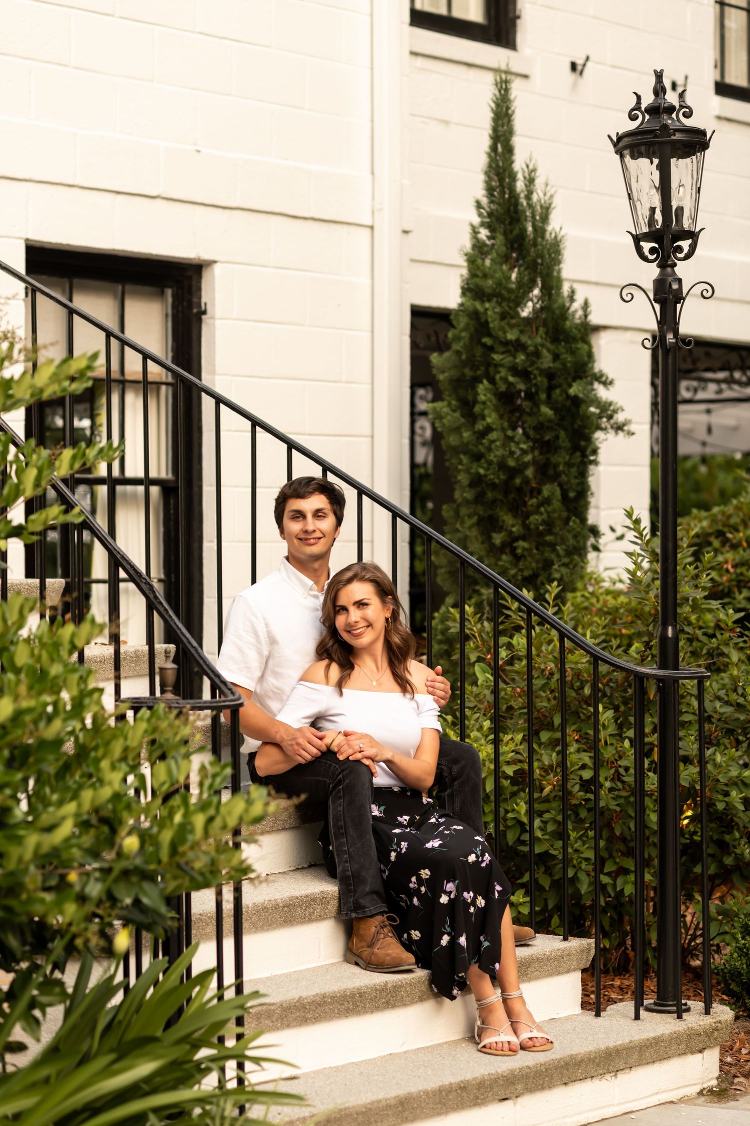 A couple sits together on white steps outside an elegant home with wrought iron railings and ornate lamp post.