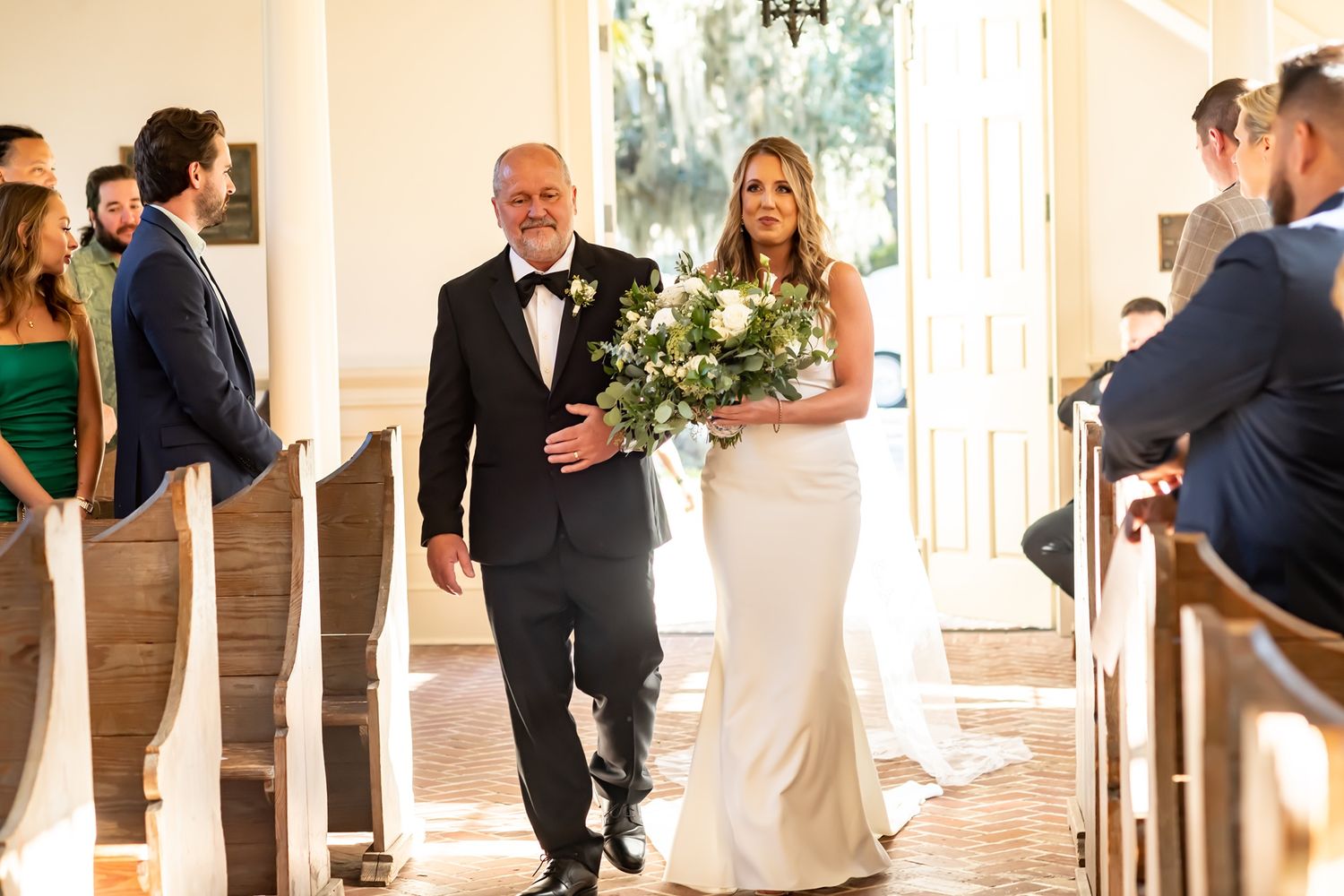 A person in formal attire escorts someone wearing an elegant white wedding gown down a church aisle.