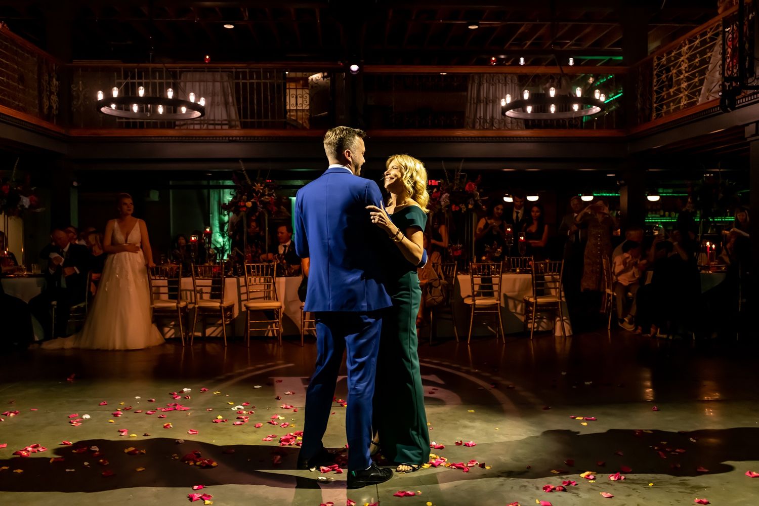 Two people share a dance on an elegant ballroom floor with rose petals scattered around them as guests watch from tables.