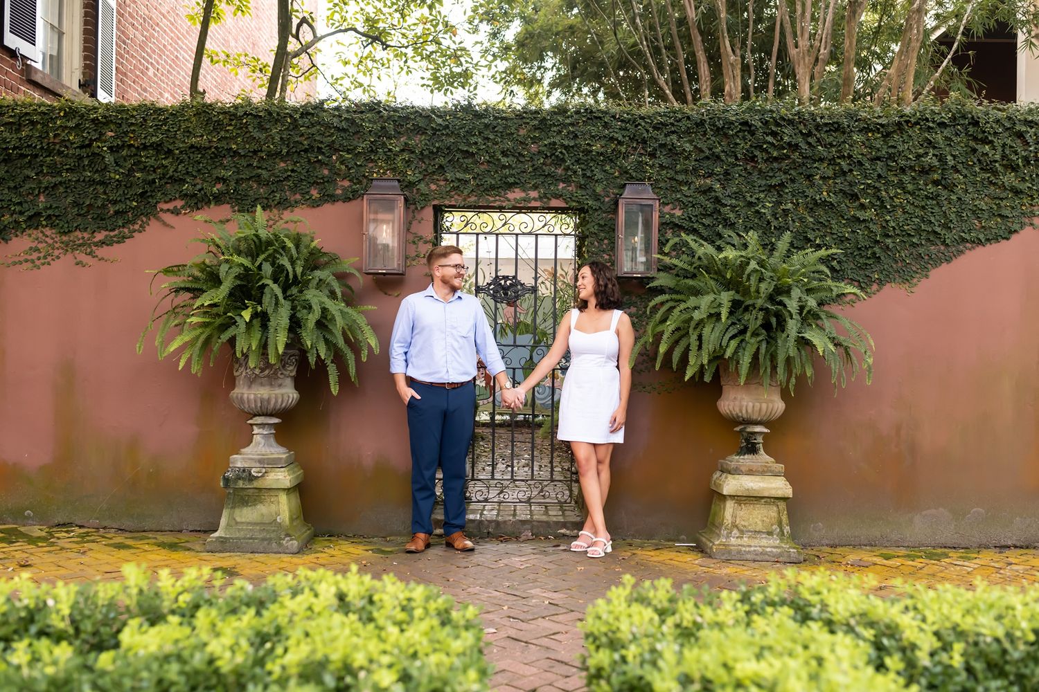 A garden walkway lined with lush greenery and decorative stone urns leads to a terracotta-colored Mediterranean villa.