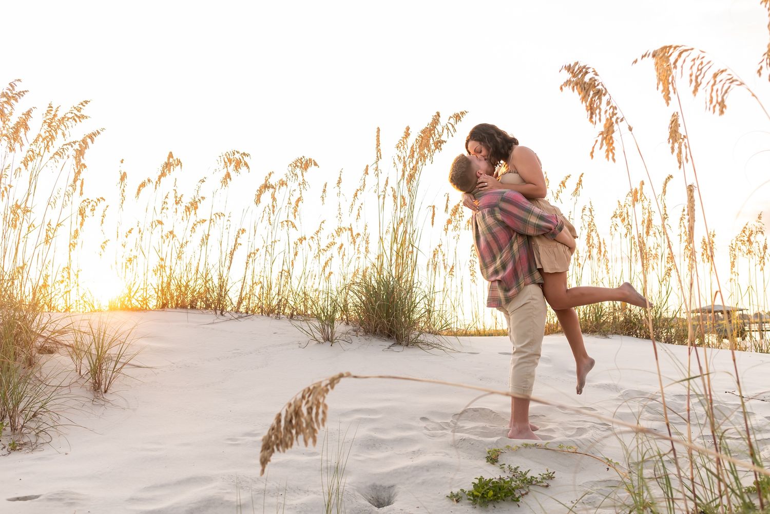 Romantic couple embraces on sandy beach with golden sunlight streaming through sea oats at sunset.