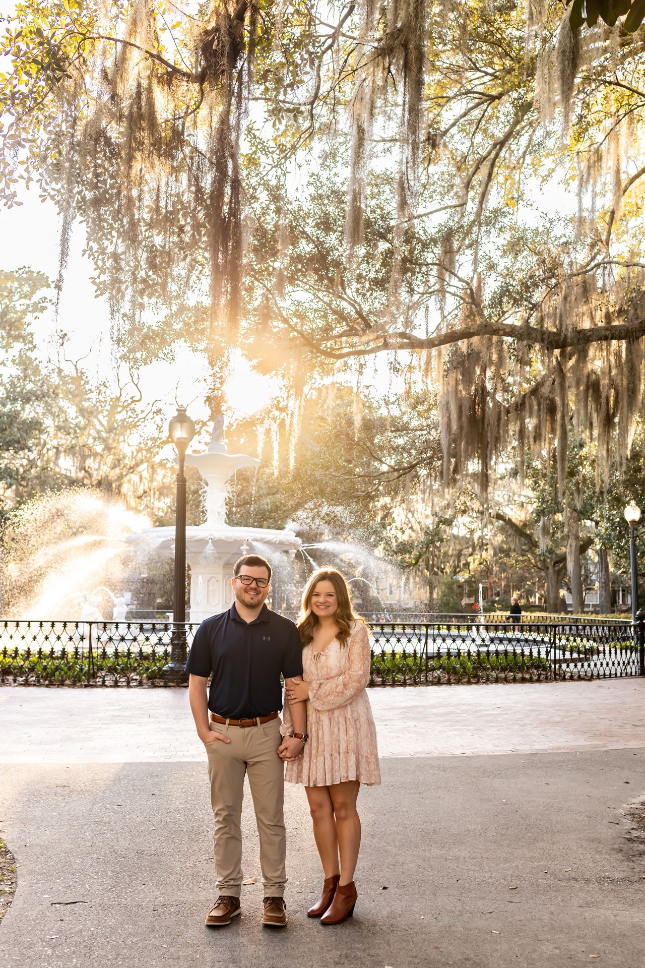 A couple stands on a path beneath Spanish moss-draped trees with golden sunlight filtering through at sunset.