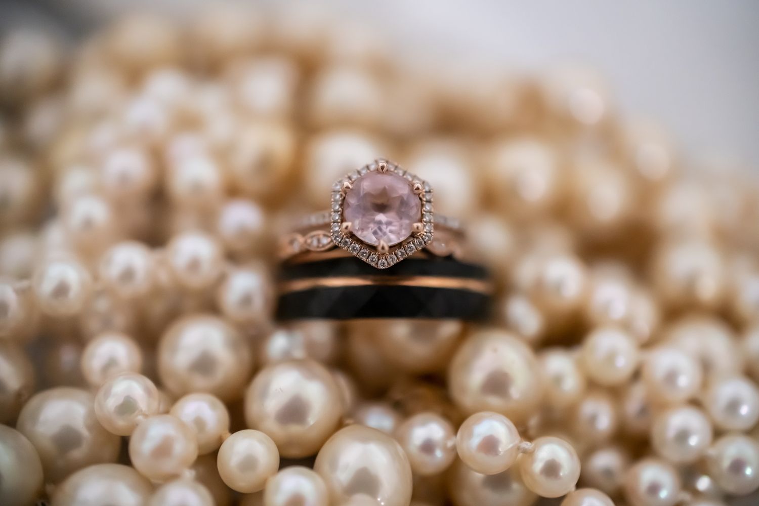 Rose gold engagement ring with pink center stone rests elegantly on white pearl necklace in soft focus.