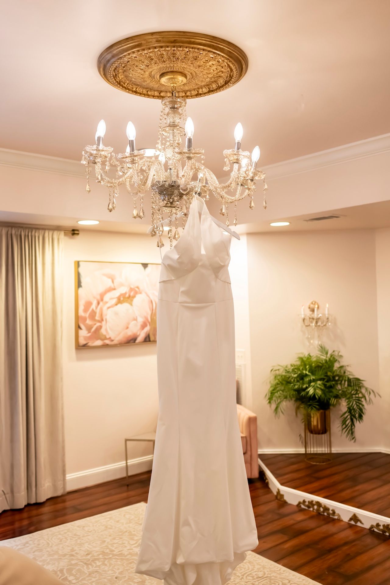 A white wedding dress hangs elegantly from an ornate crystal chandelier in a room with hardwood floors and cream walls.