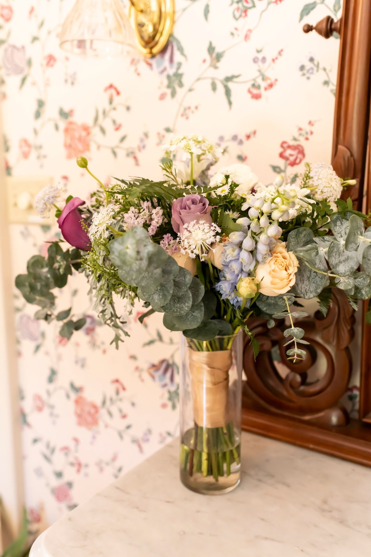 A romantic floral bouquet with eucalyptus, roses, and mixed flowers in a glass vase against vintage floral wallpaper.