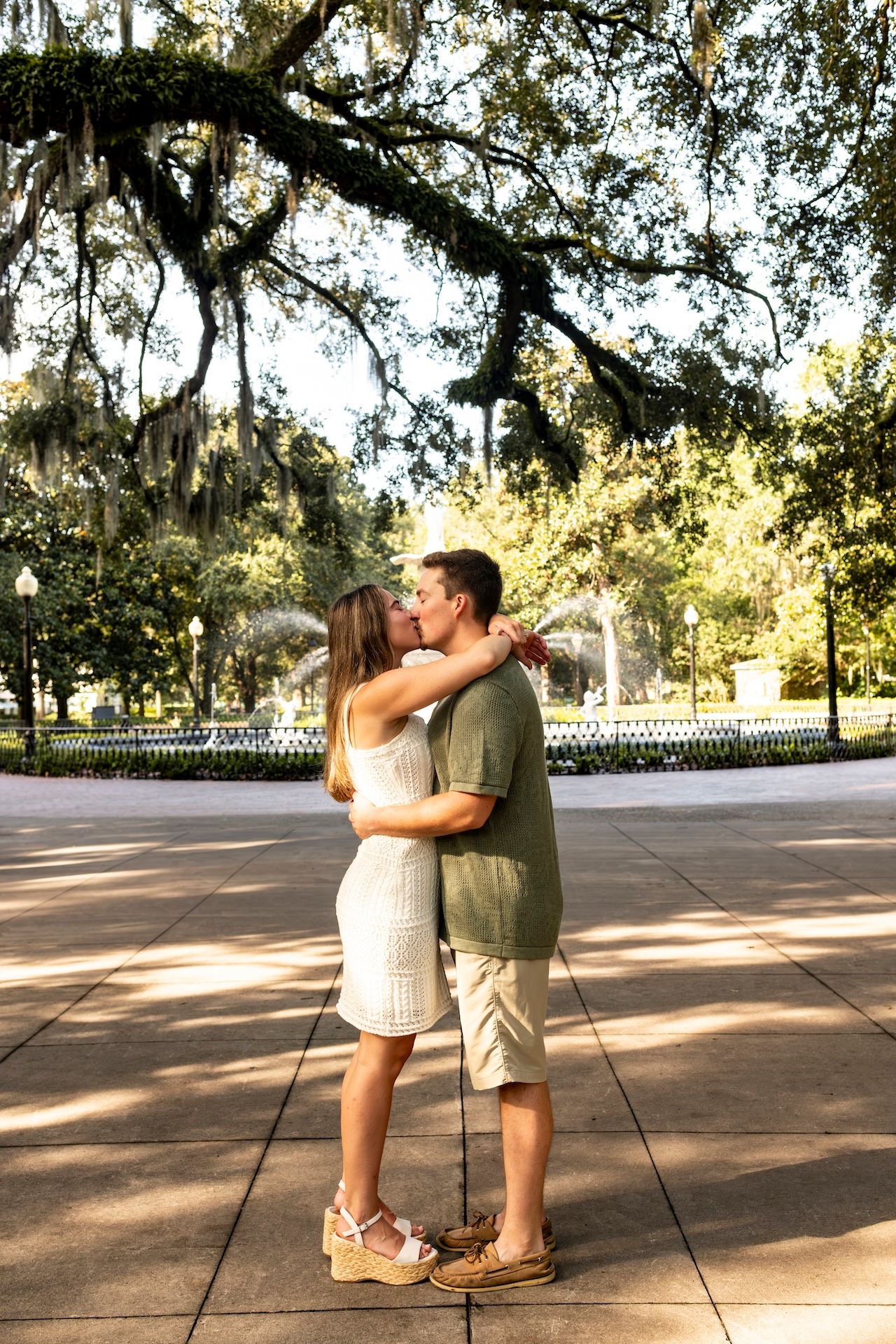 A couple embraces in a romantic moment under Spanish moss-draped trees in a scenic Southern park setting.