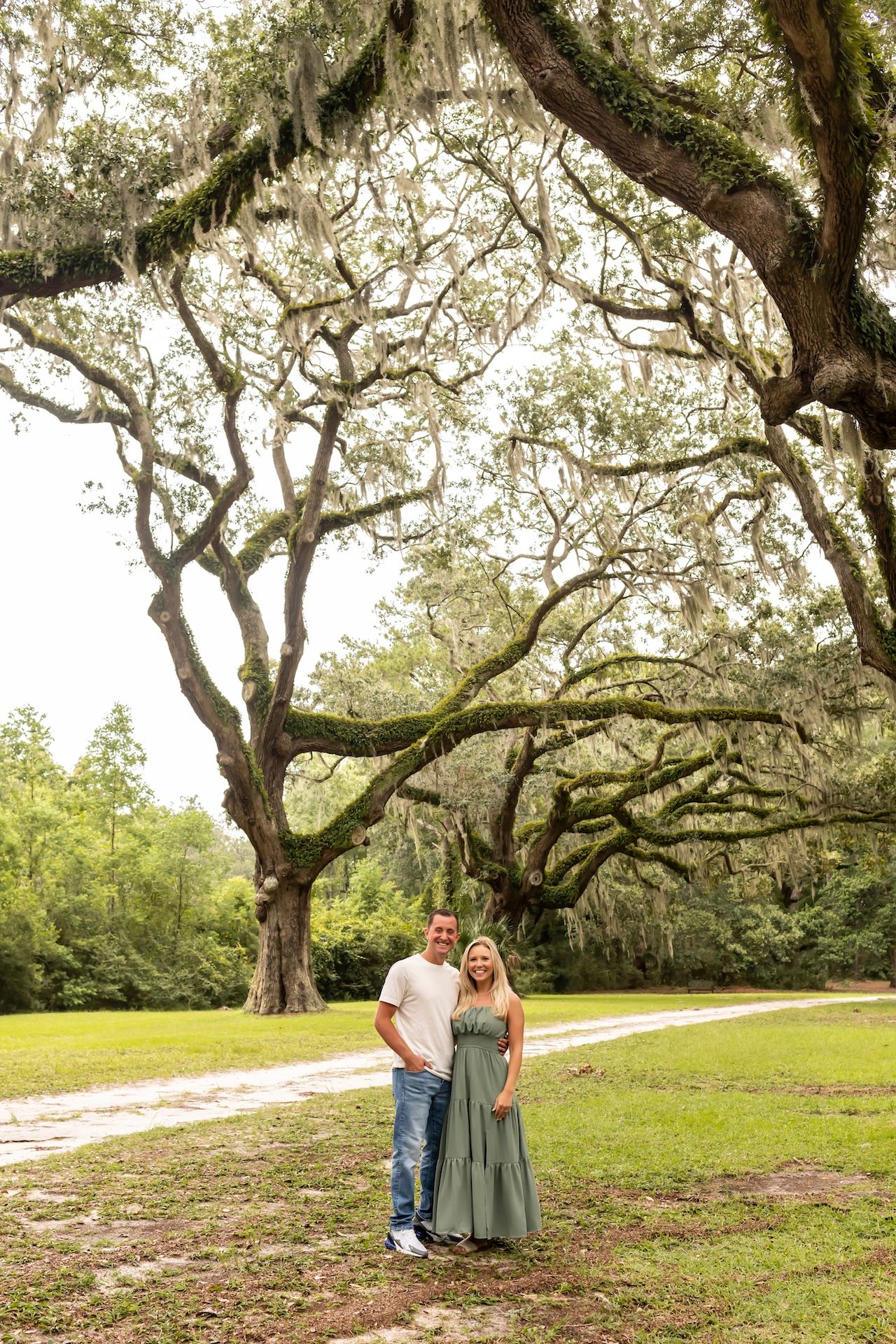 A couple stands beneath towering live oak trees draped in Spanish moss on a grassy path in a Southern garden setting.