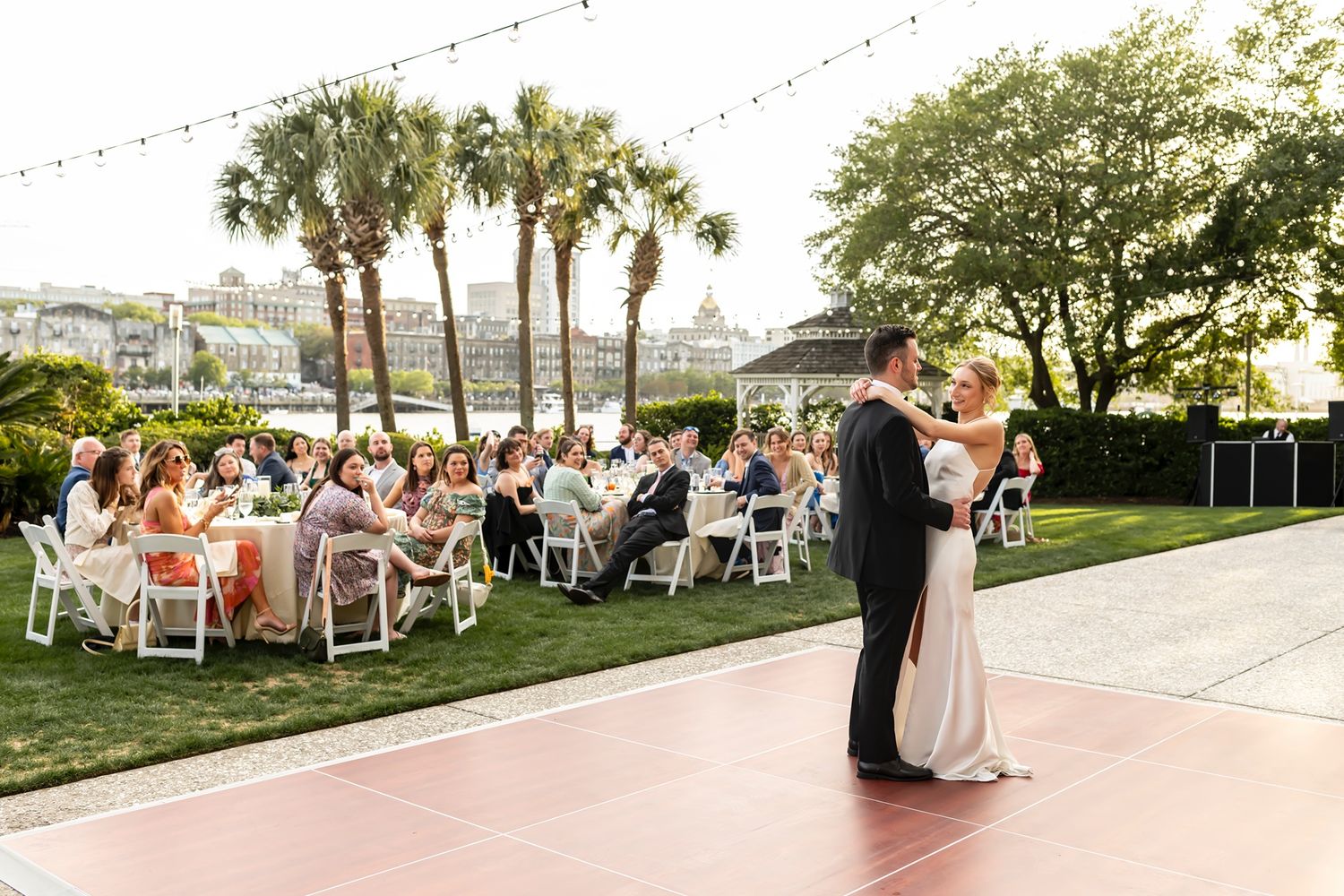 Couple shares their first dance on outdoor terrace while wedding guests watch from lawn seating at sunset.