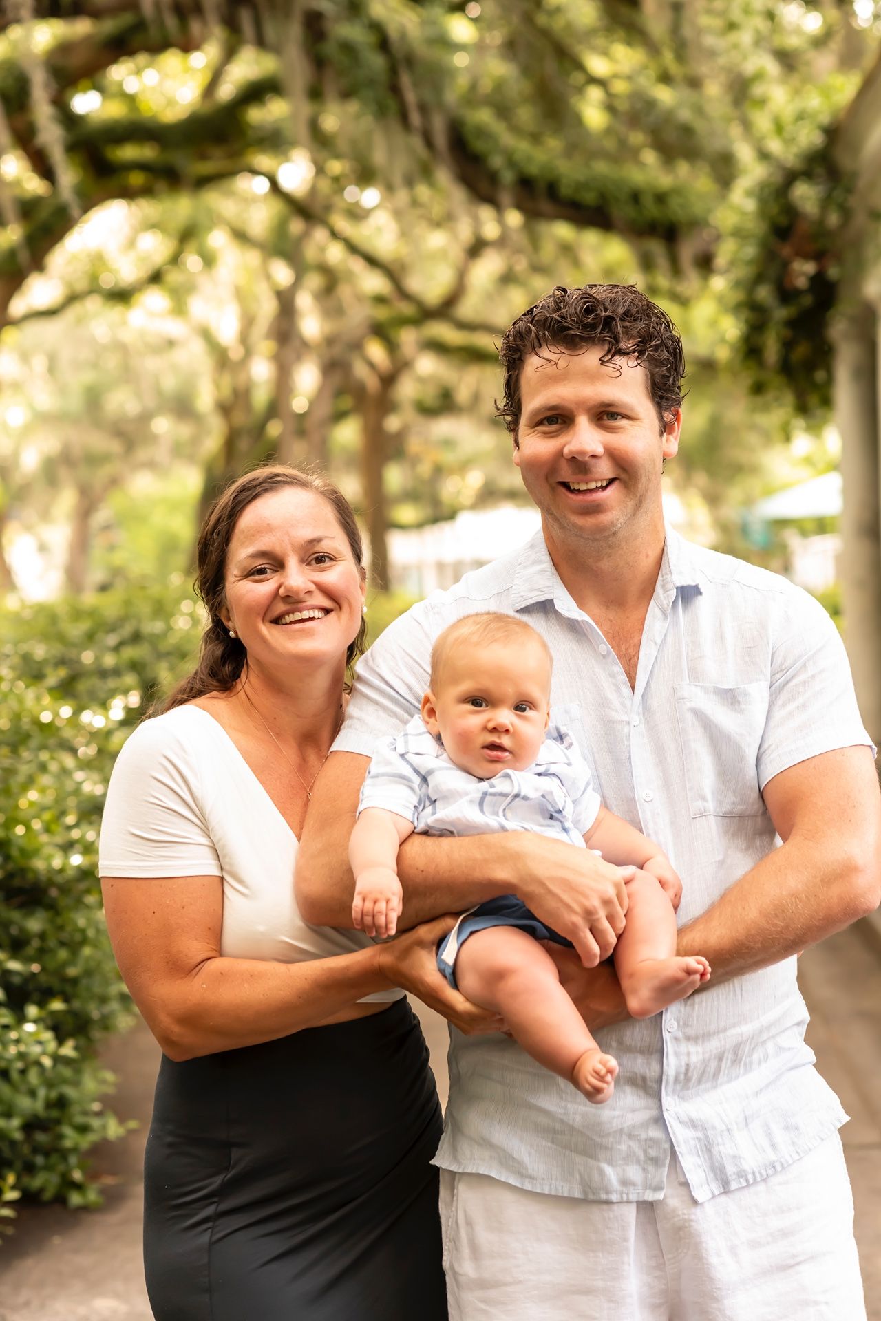 A family poses outdoors in a garden setting with tall trees creating a natural backdrop on a sunny day.