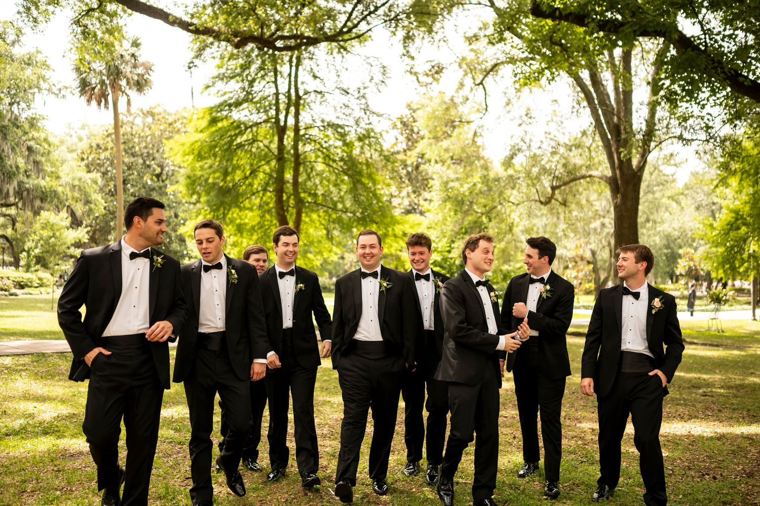 A group of groomsmen in black tuxedos stand together in a relaxed pose outdoors in a sunlit park setting.
