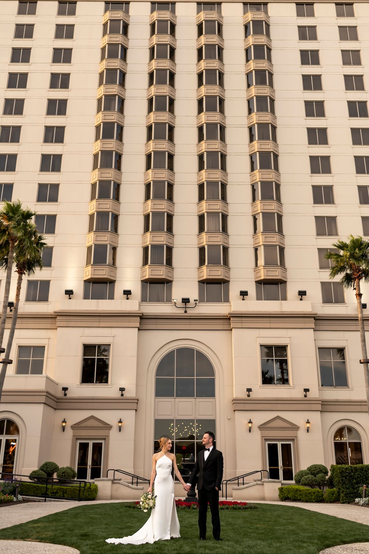 Bride and groom standing together on green lawn in front of elegant tall hotel building with palm trees.