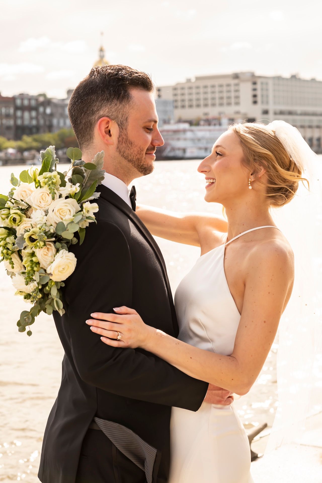 Bride and groom embrace near waterfront, she holds white floral bouquet, wearing wedding gown and tuxedo.