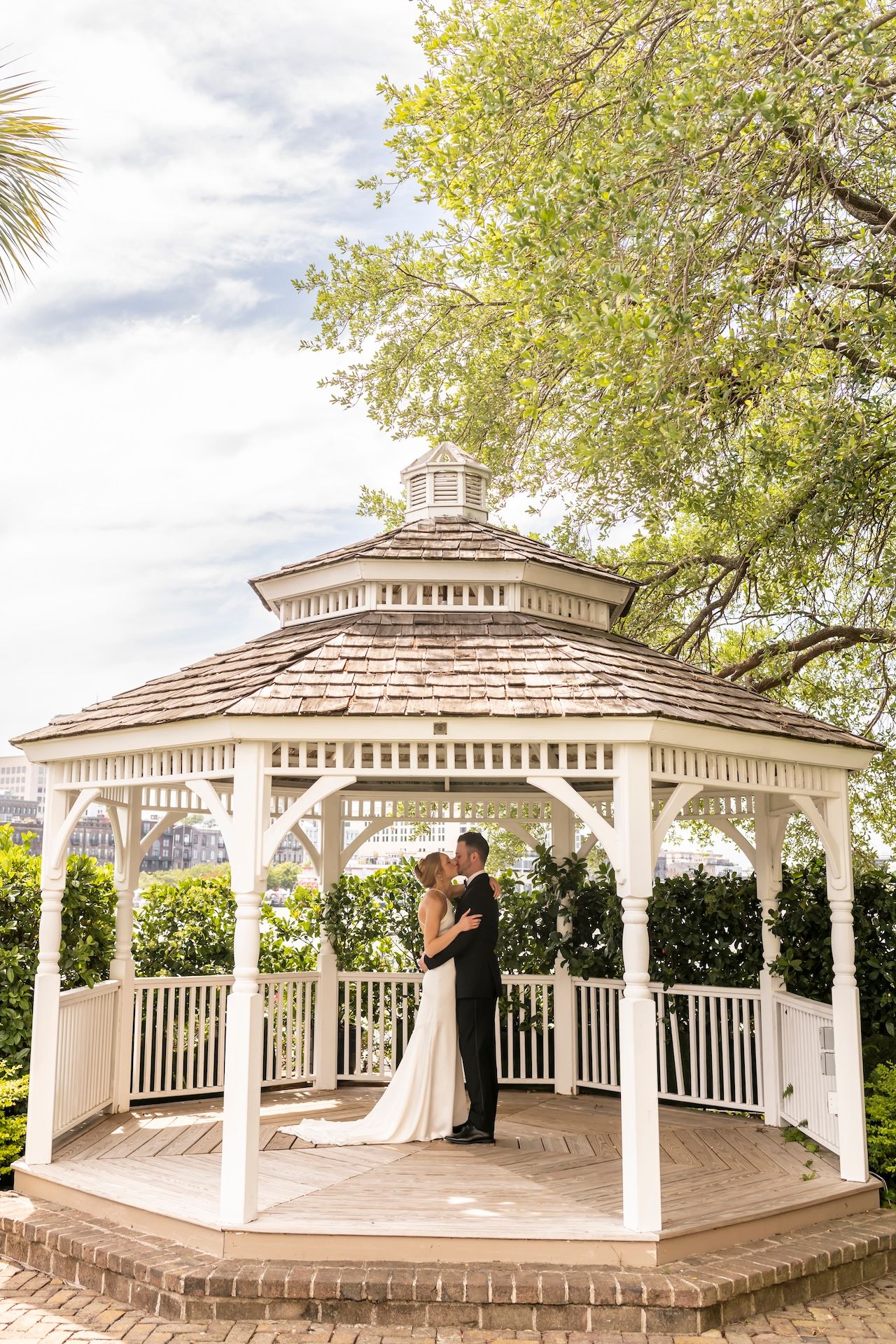 Bride and groom share romantic embrace inside white gazebo surrounded by lush garden greenery.