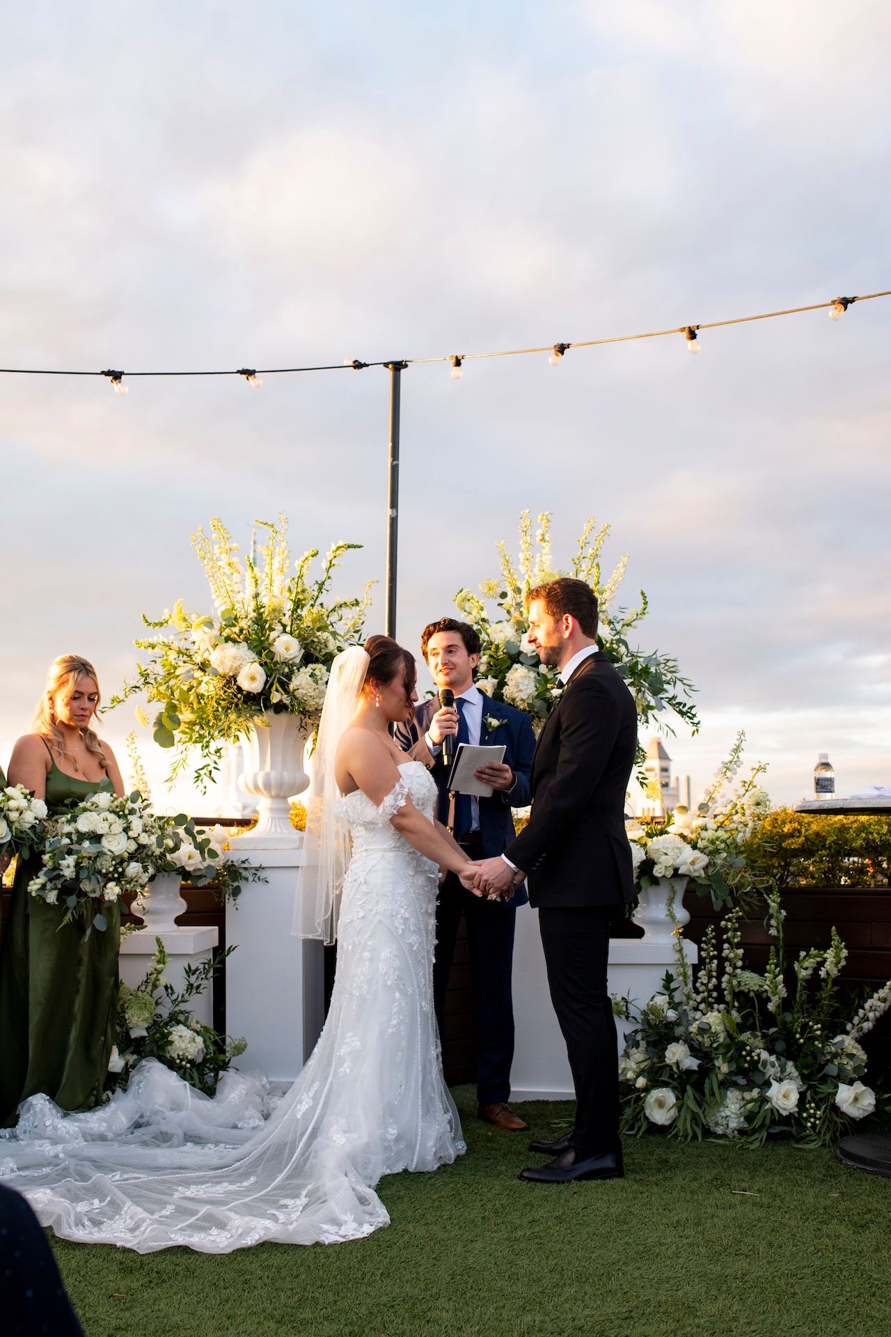 Outdoor wedding ceremony at sunset with string lights and white floral arrangements on a grassy lawn by the ocean.