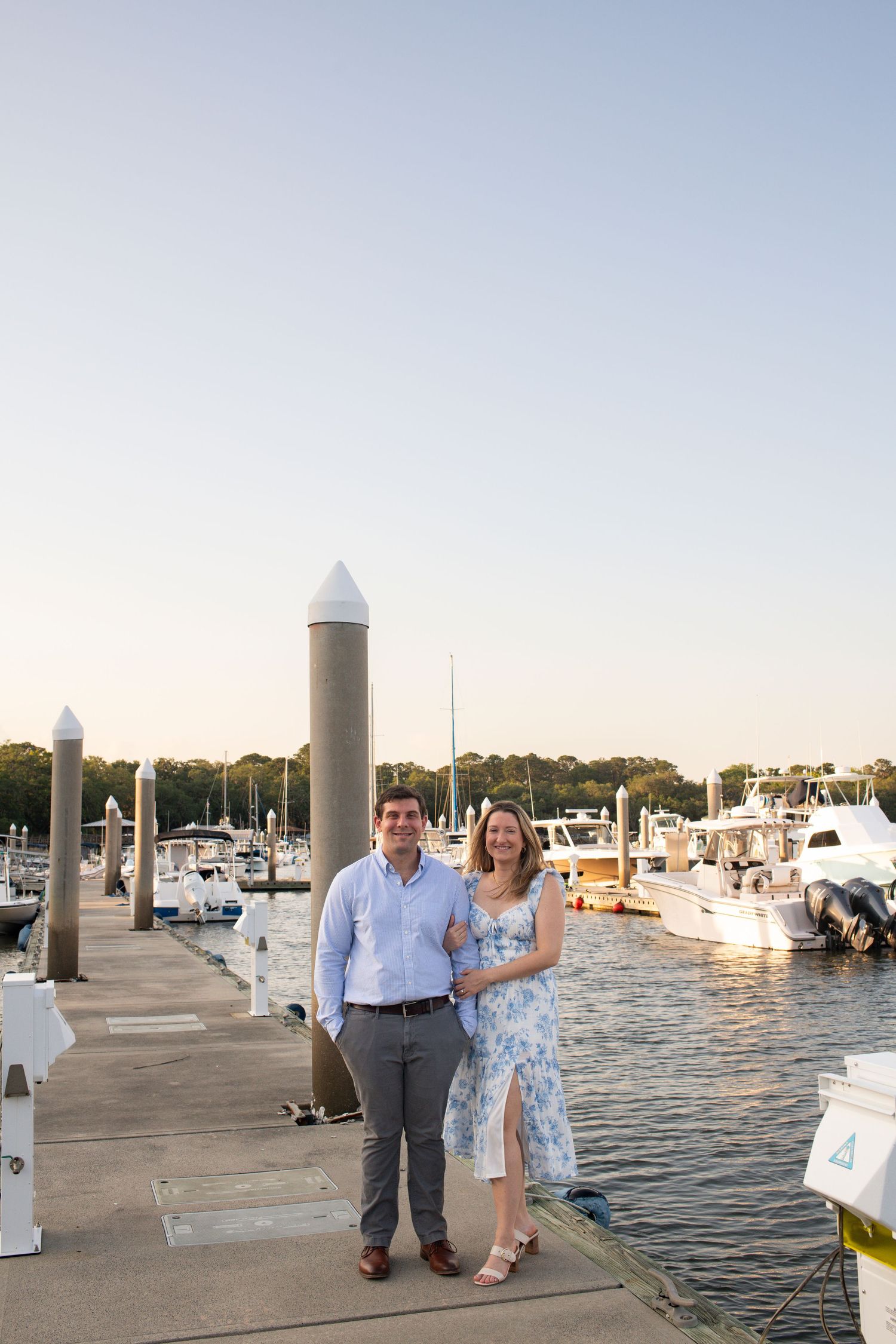 Couple enjoying a romantic evening stroll along a marina dock with boats and sunset views in the background.