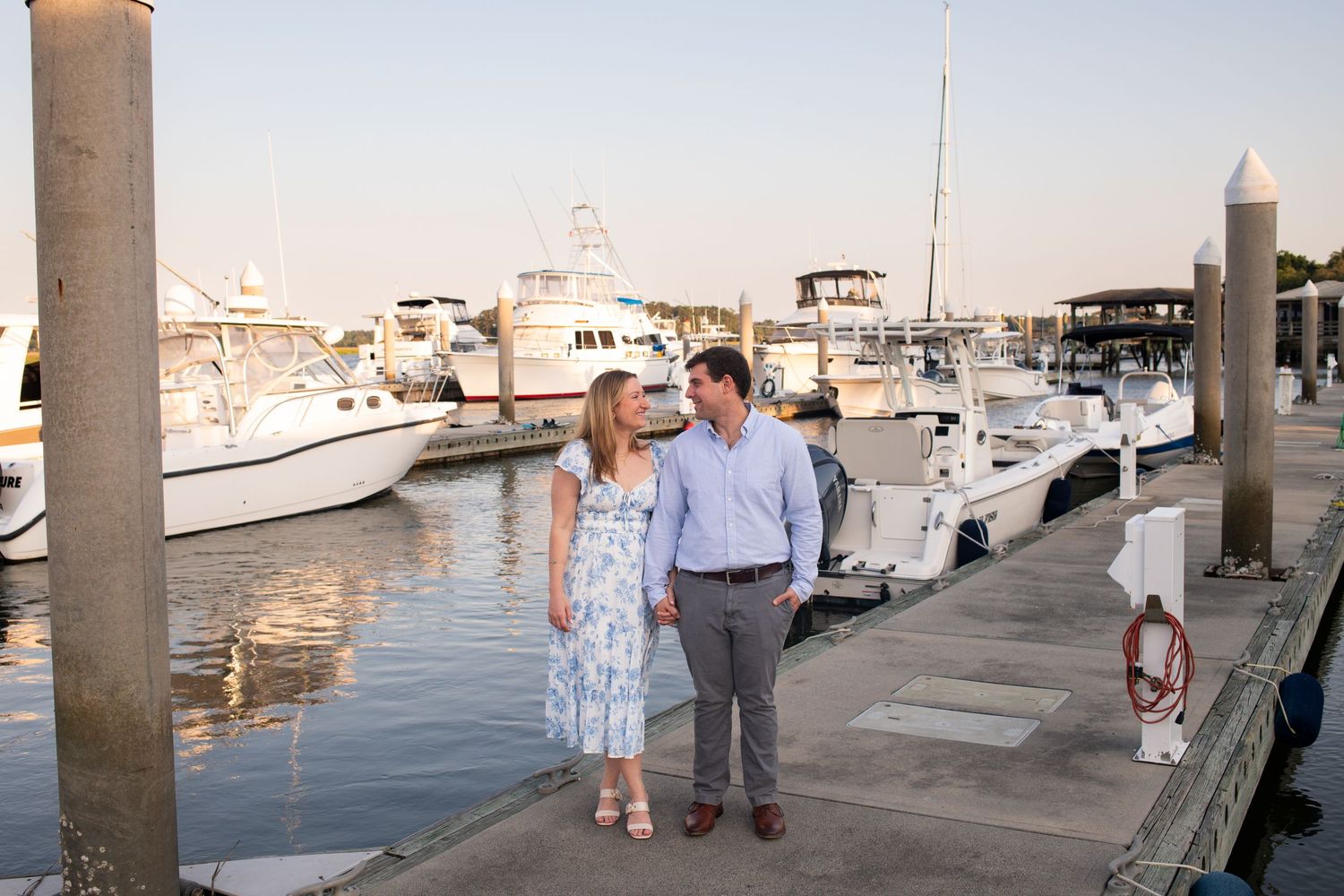 People walking along a marina dock with boats and buildings visible in the background during sunset.