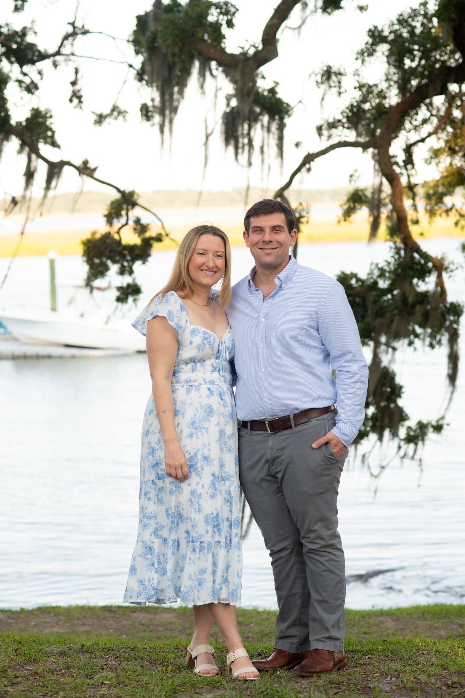 A couple poses together outdoors near a waterfront with Spanish moss hanging from trees during sunset.