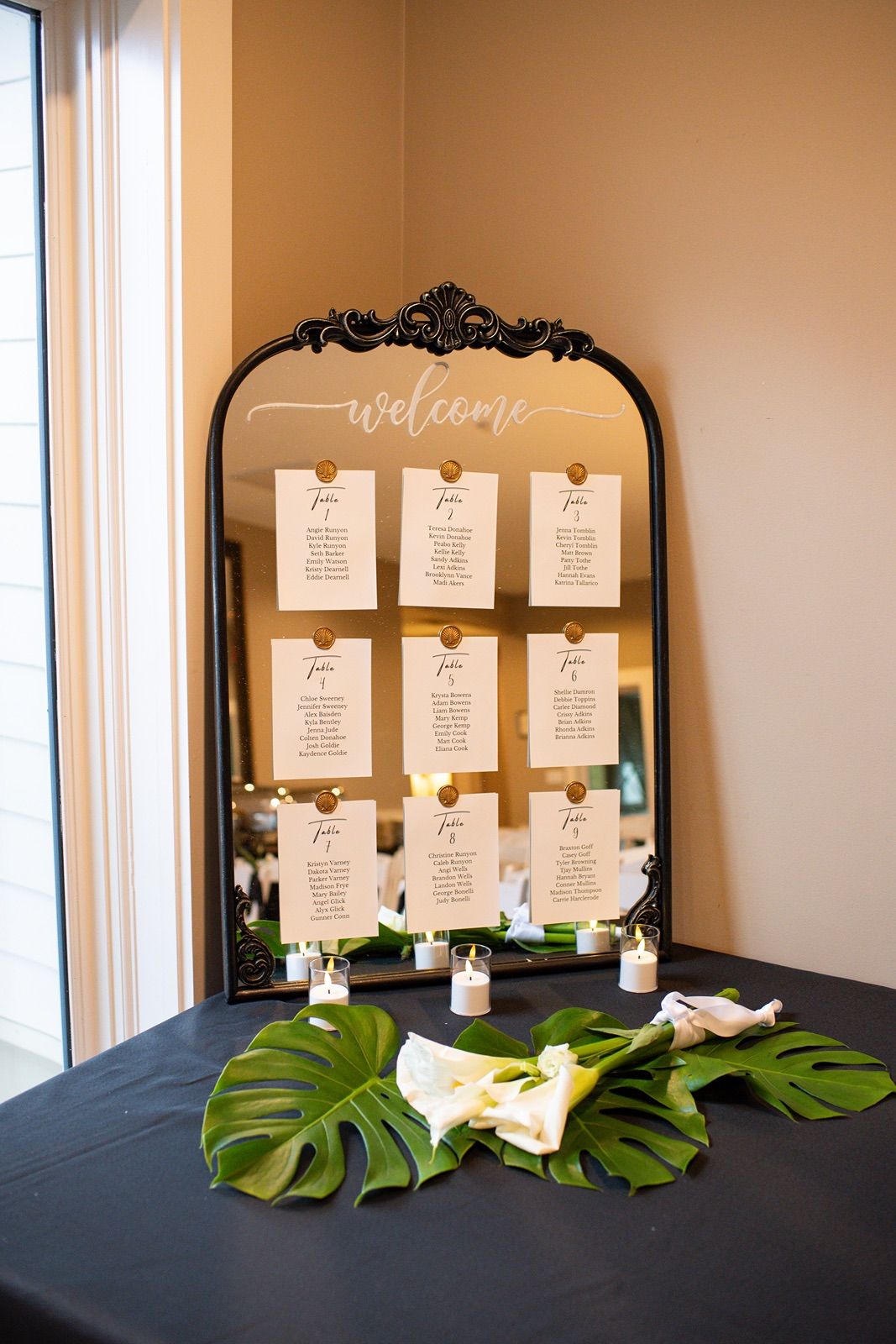 Elegant black framed seating chart display with tropical monstera leaves and white cards.