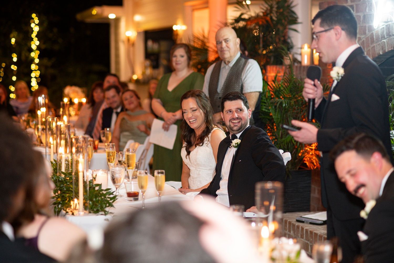 Bride and groom smile during wedding reception toast at candlelit head table surrounded by guests.