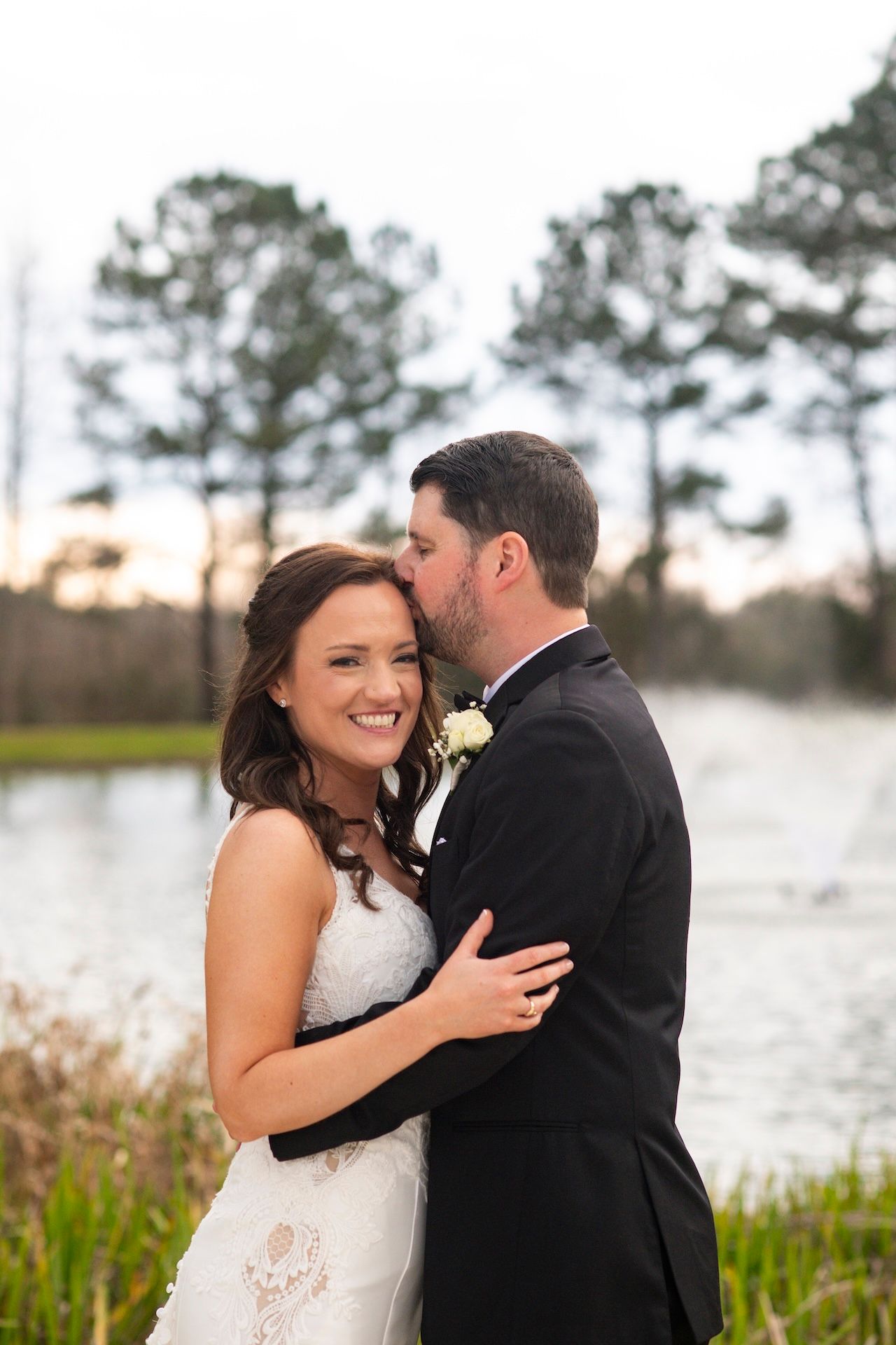 Bride and groom embrace near a serene pond at sunset, with the groom kissing her cheek as she smiles.