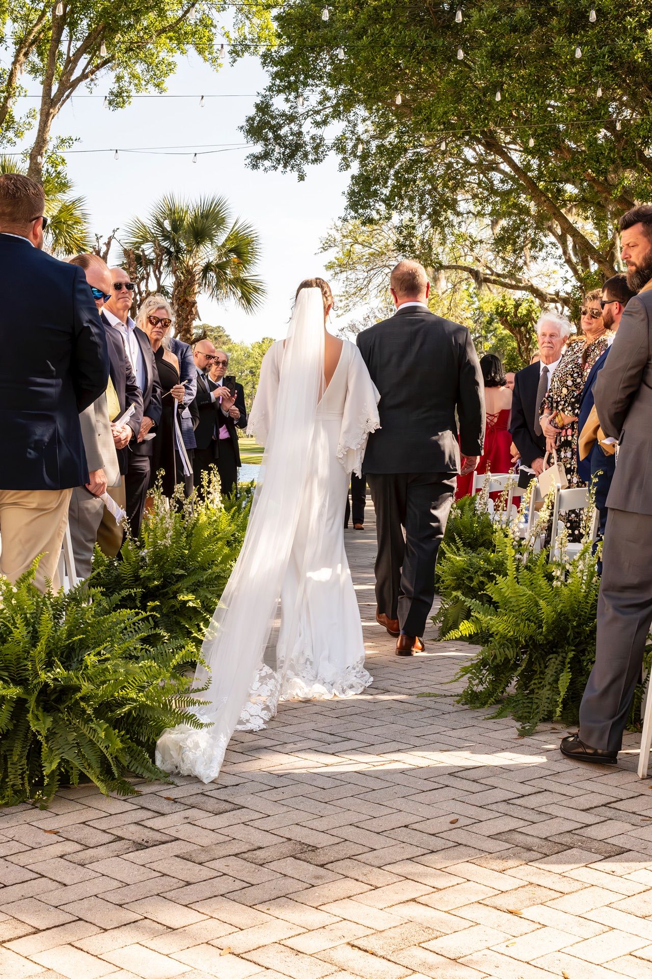 Wedding guests and bridal party gather on a stone patio surrounded by lush greenery and palm trees.