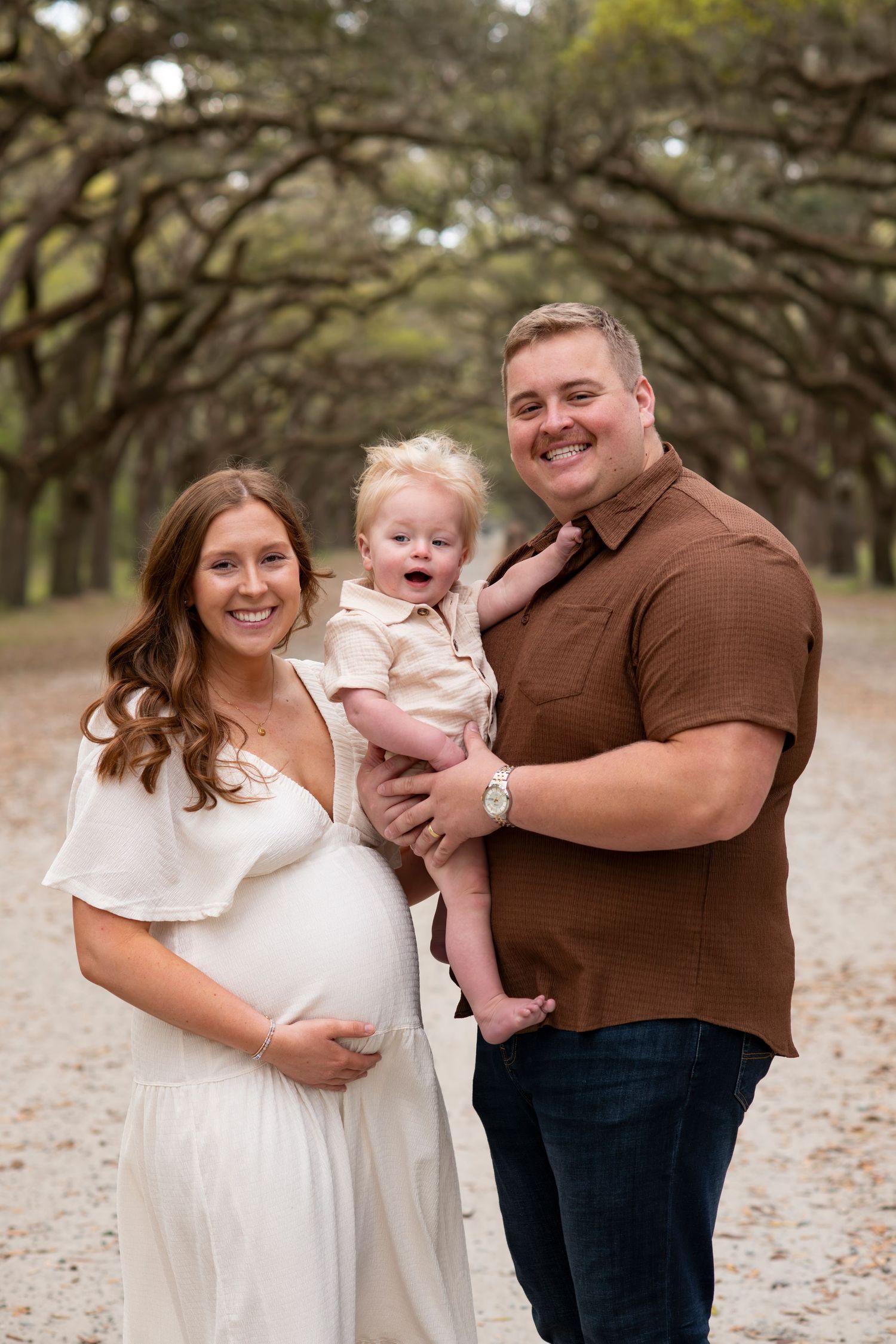 Pregnant woman in white dress and man in brown shirt hold toddler on tree-lined path for maternity photo.