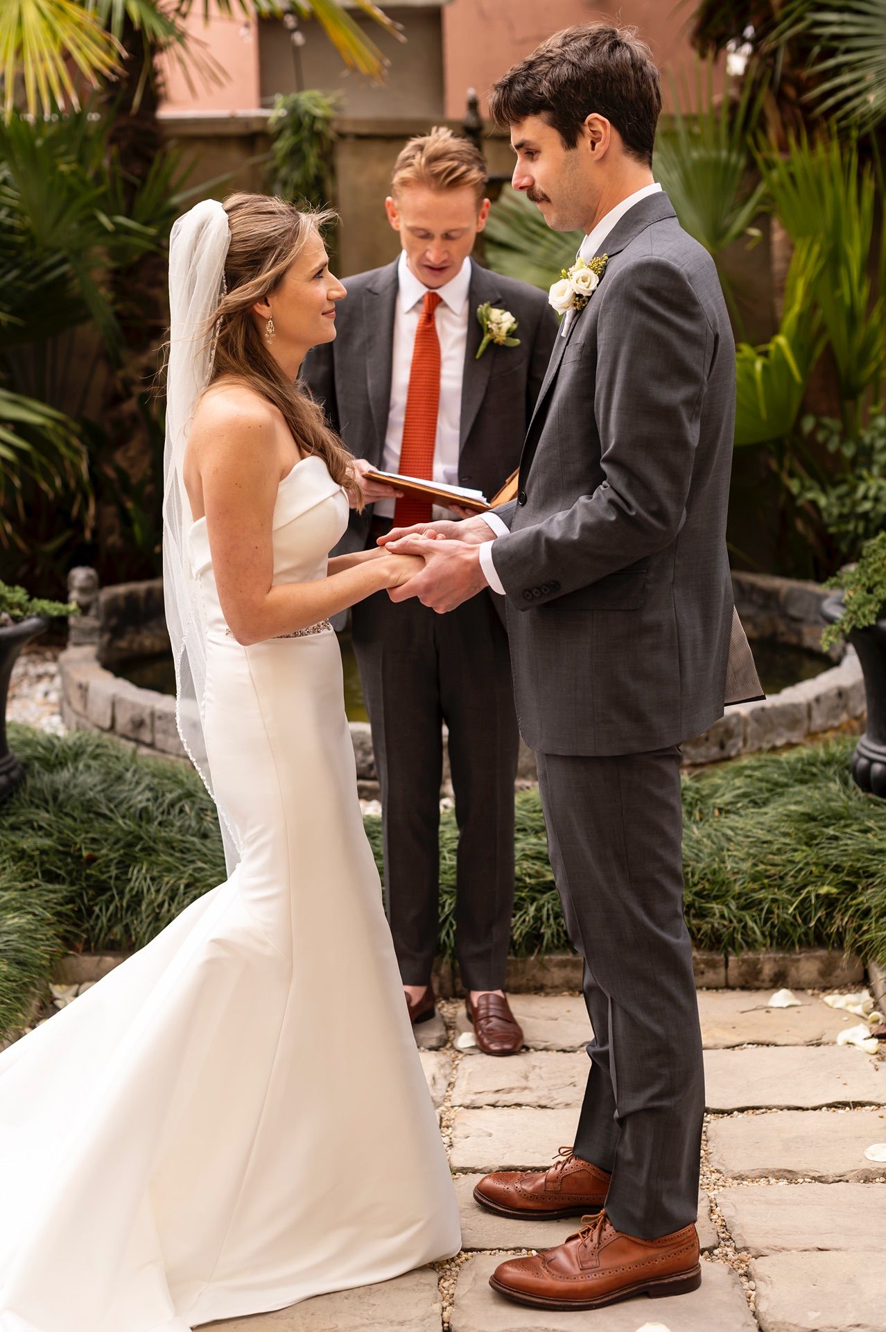 Bride in white gown exchanges rings with groom during outdoor wedding ceremony with palm trees in background.