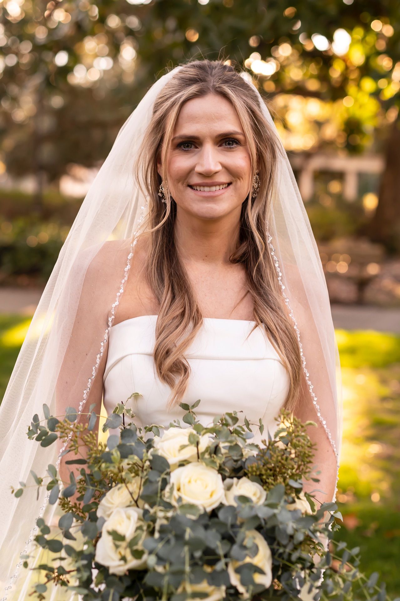 Bride in white strapless gown smiles while holding eucalyptus bouquet during golden hour outdoor wedding photoshoot.