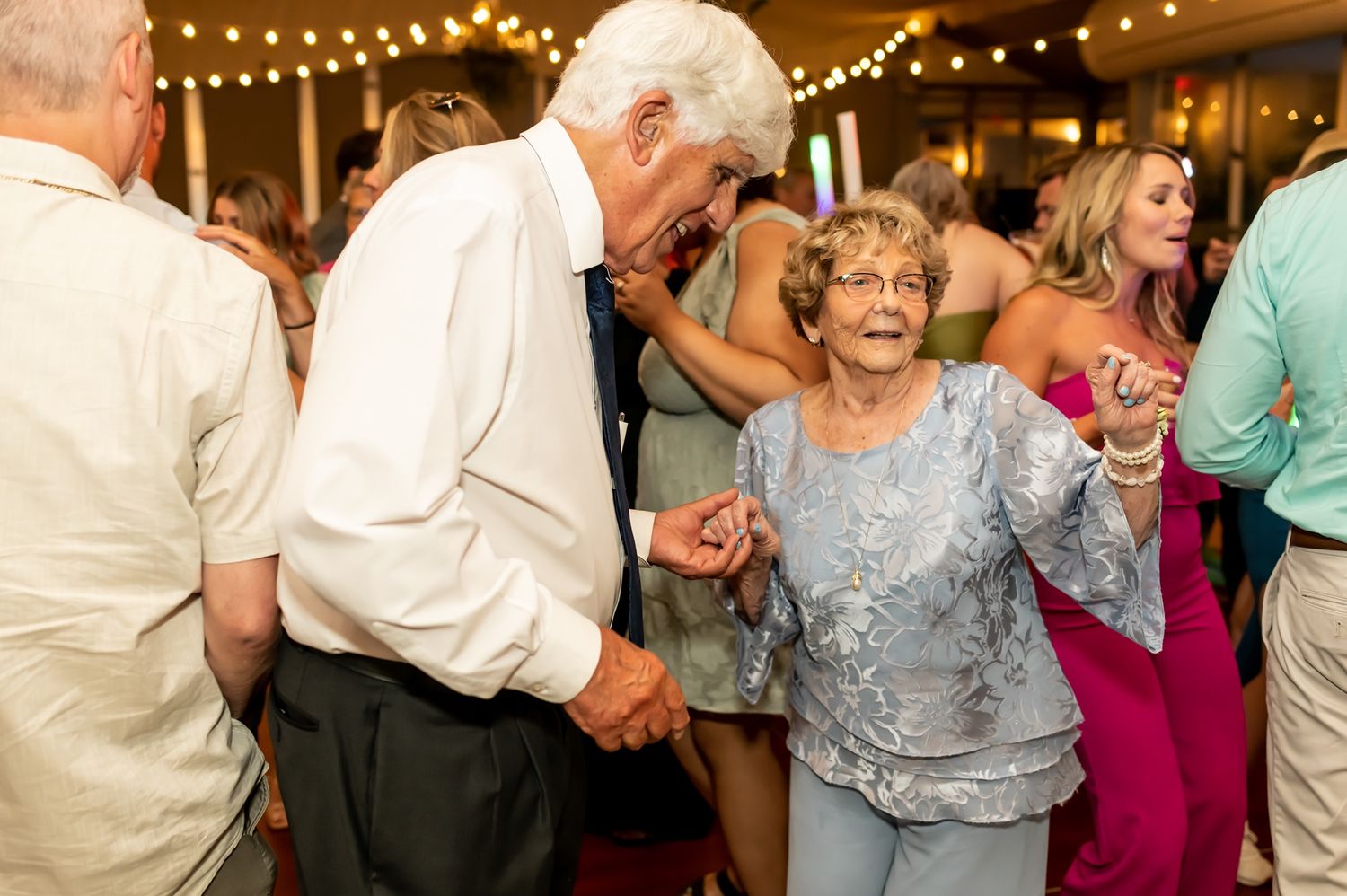 Elderly couple dancing together at a reception venue decorated with string lights while others celebrate around them.