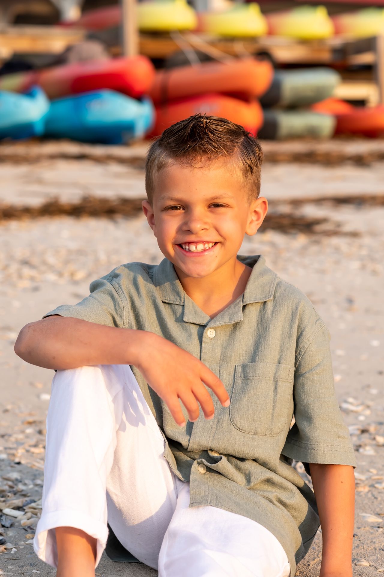 Young child in sage green shirt and white pants sits casually outdoors with colorful boats in the background.