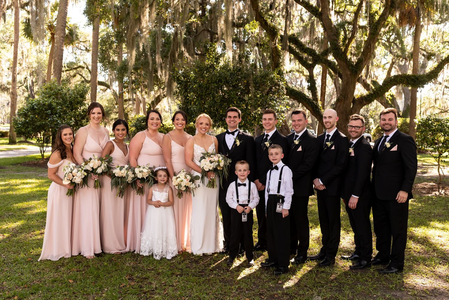 Wedding party posed outdoors beneath Spanish moss-draped oak trees wearing blush pink gowns and black tuxedos.